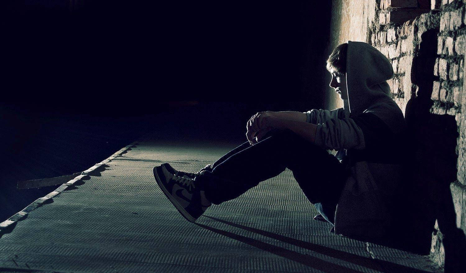 Alone Boy 桌布 壁紙 — 一個孤獨嘅人著住連帽衫，喺夜晚昏暗嘅小巷入面挨住磚牆坐低，被一道光照射住。