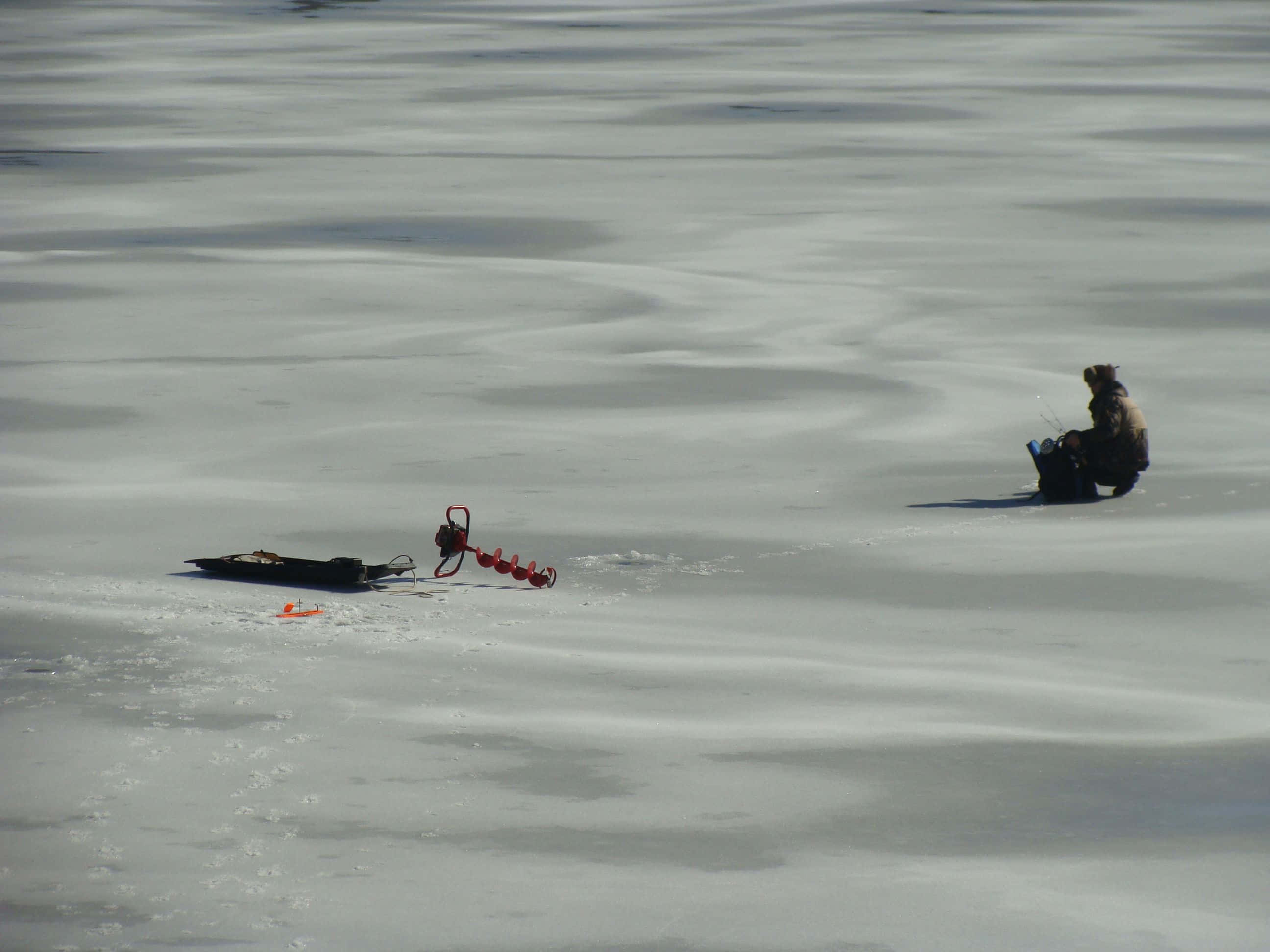 Ice Fishing 桌布 壁紙 — 一個孤獨嘅冰釣者坐喺結冰嘅湖面上，附近有個紅色冰鑽，喺明亮嘅日光下。
