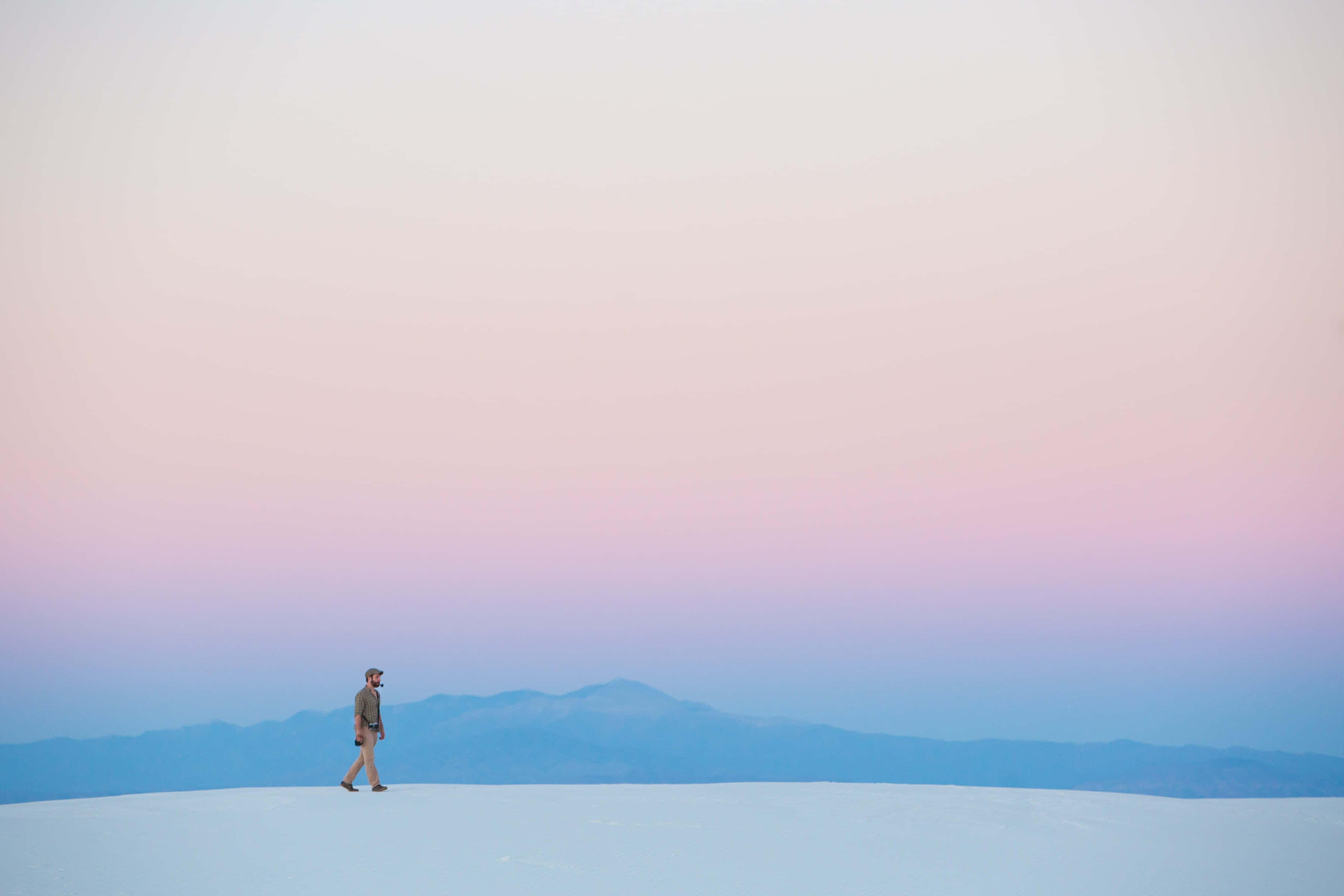 Minimalism 桌布 壁紙 — 一個孤獨的人在一片廣闊的白色沙漠中行走，天空是柔和的粉紅色和藍色暮色，遠處有山脈。