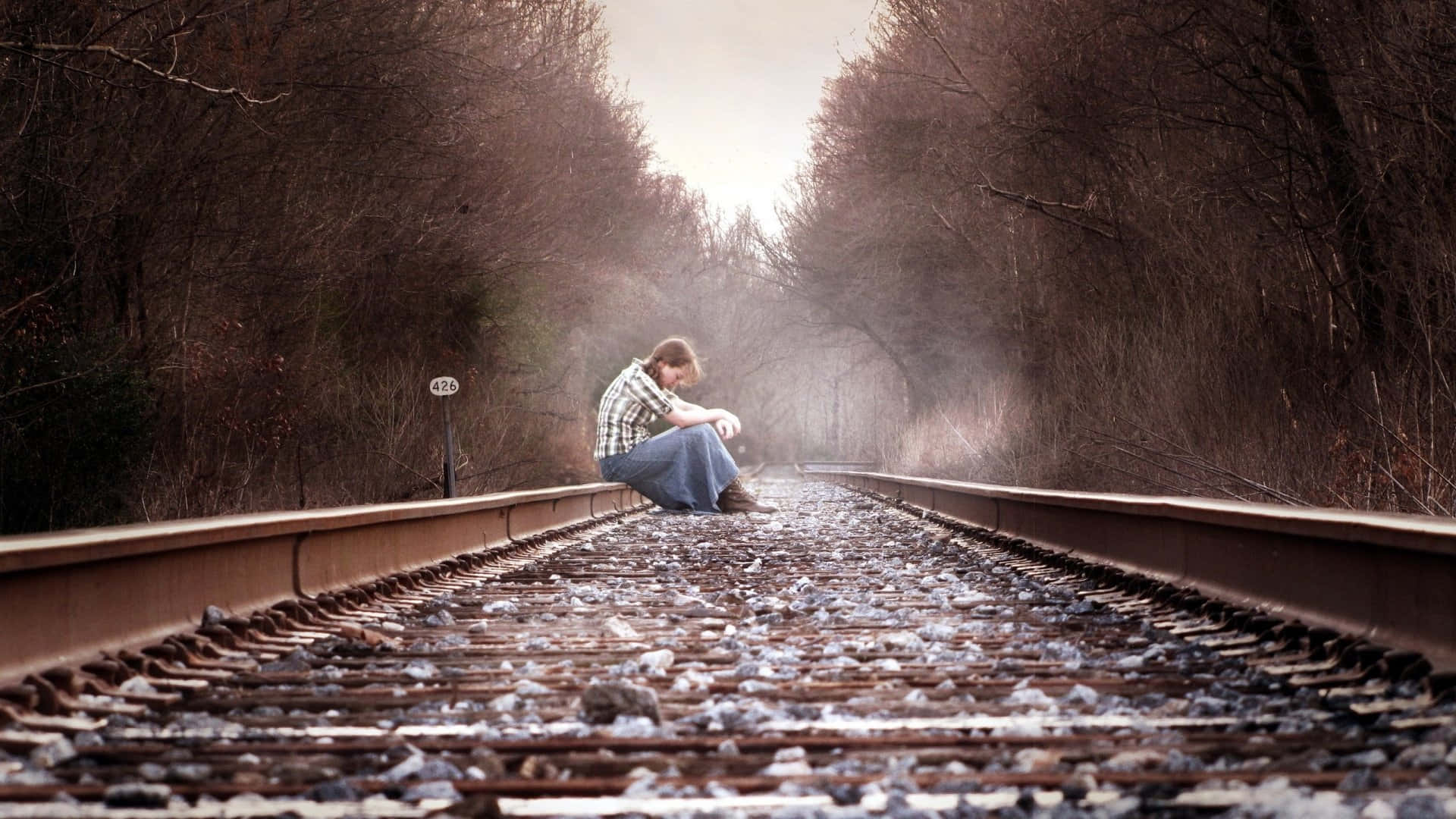 sadness 桌布 壁紙 — 一個孤獨的人坐在生鏽的火車軌道上沉思，周圍是光禿禿的冬日樹木，天空朦朧。