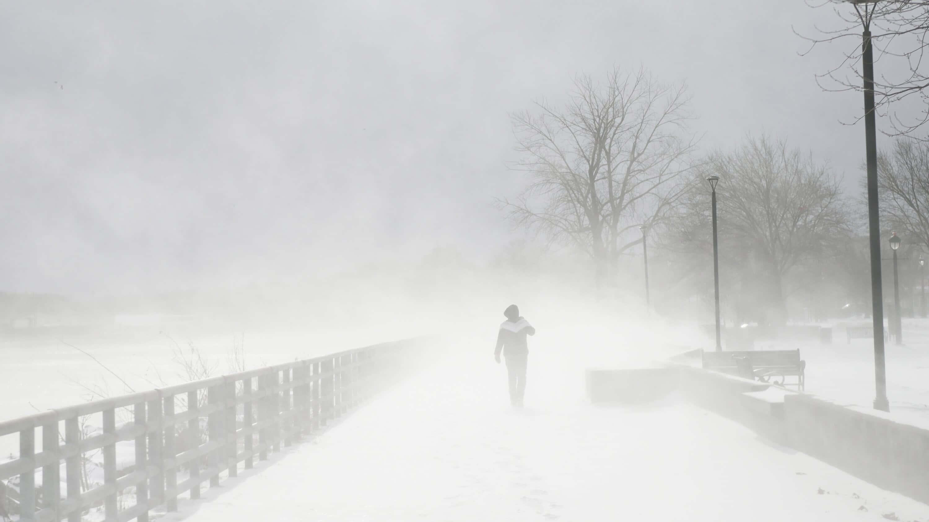 寒冷 桌布 壁紙 — 一個孤獨的人穿過暴風雪，走在雪覆蓋的小徑上，旁邊是欄杆，光禿禿的樹木和路燈在白色中幾乎看不見。