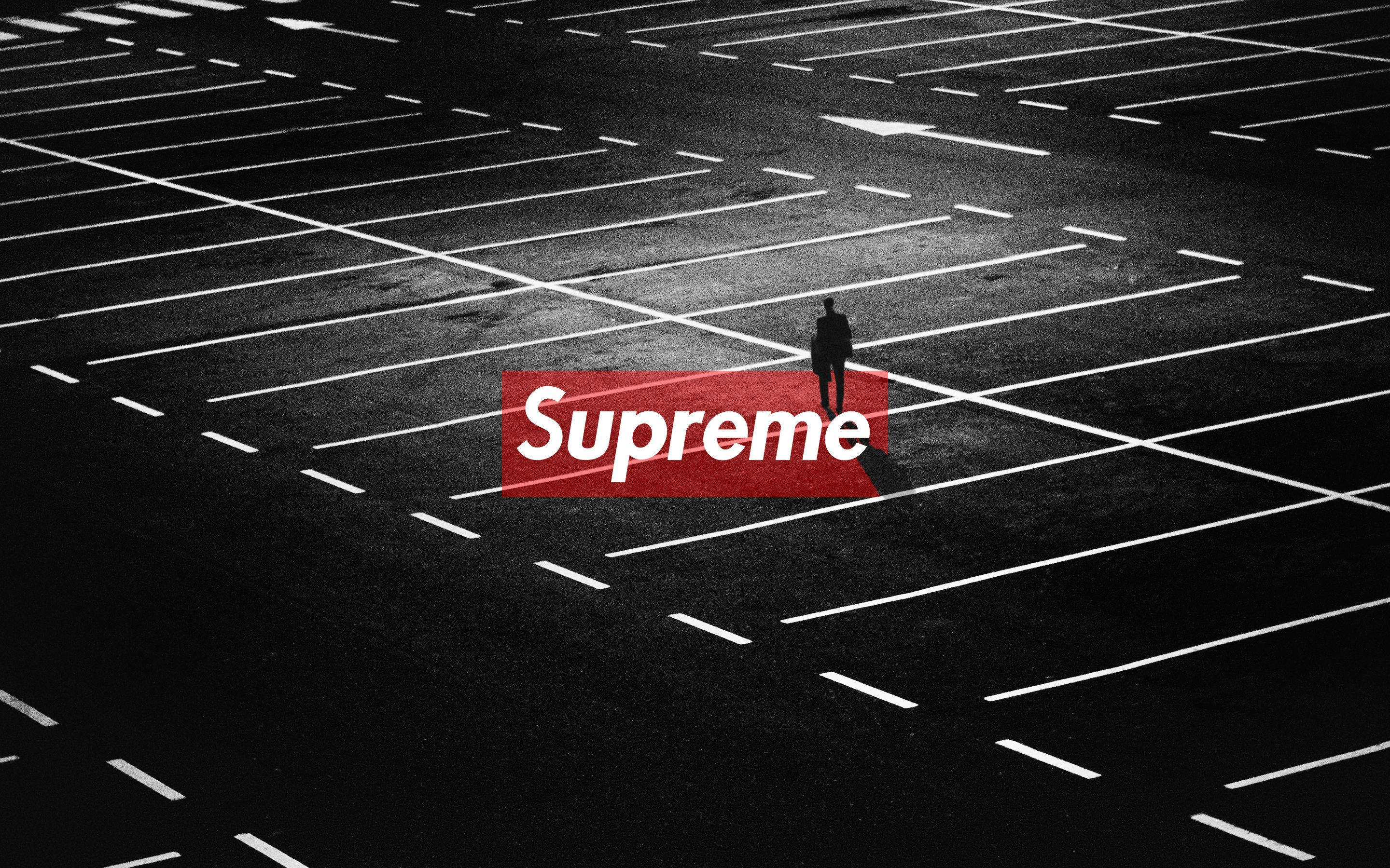 Supreme Mac 壁紙 — 一個孤獨的剪影站在廣闊的空蕩蕩的停車場中，在強烈的人造光下，帶有醒目的紅色Supreme標誌覆蓋層。