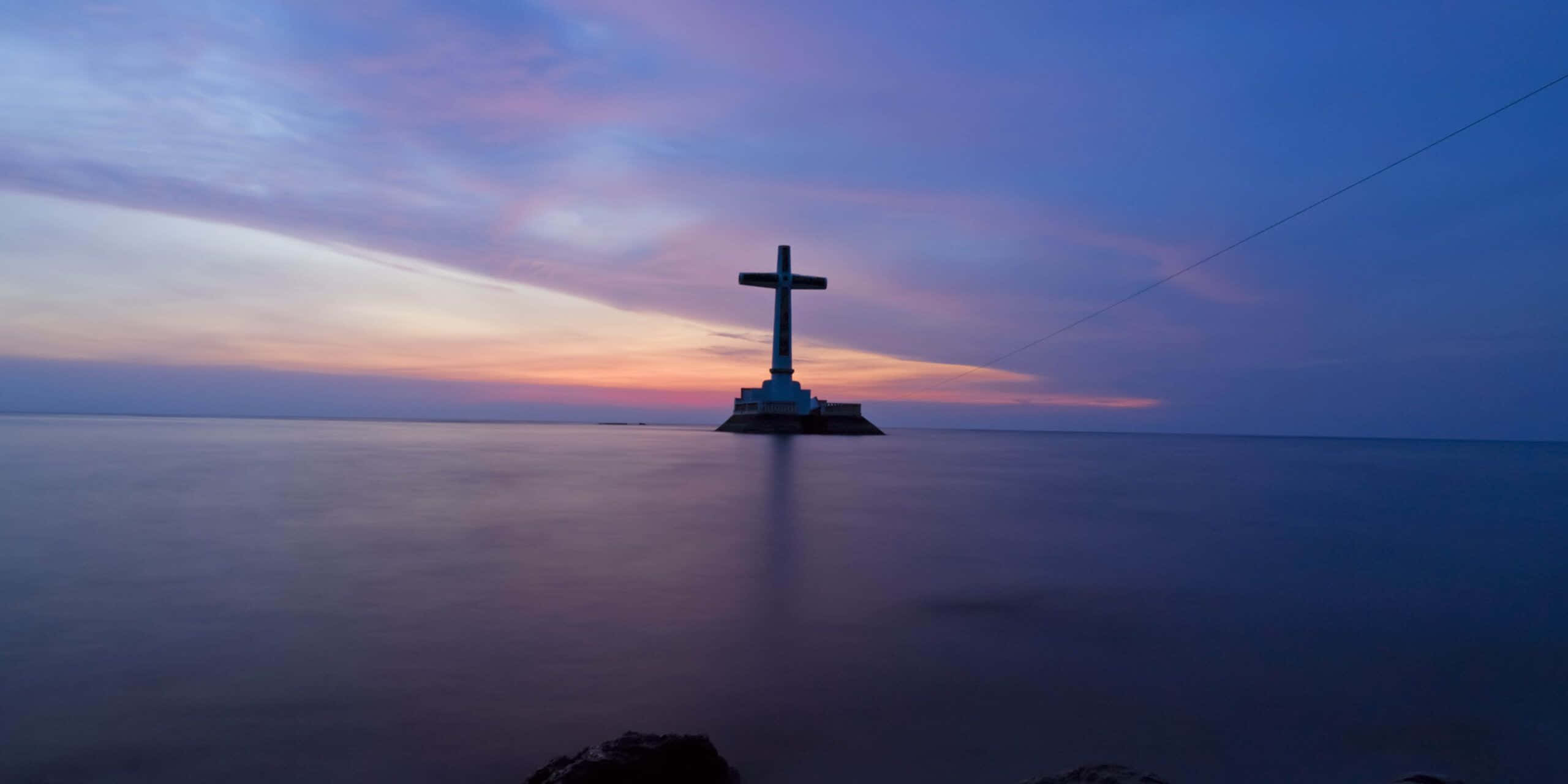 Philippines 桌布 壁紙 — 一個孤獨的十字架矗立在小島上，周圍是平靜的水面，上方是塗滿柔和紫色和粉紅色的暮色天空。