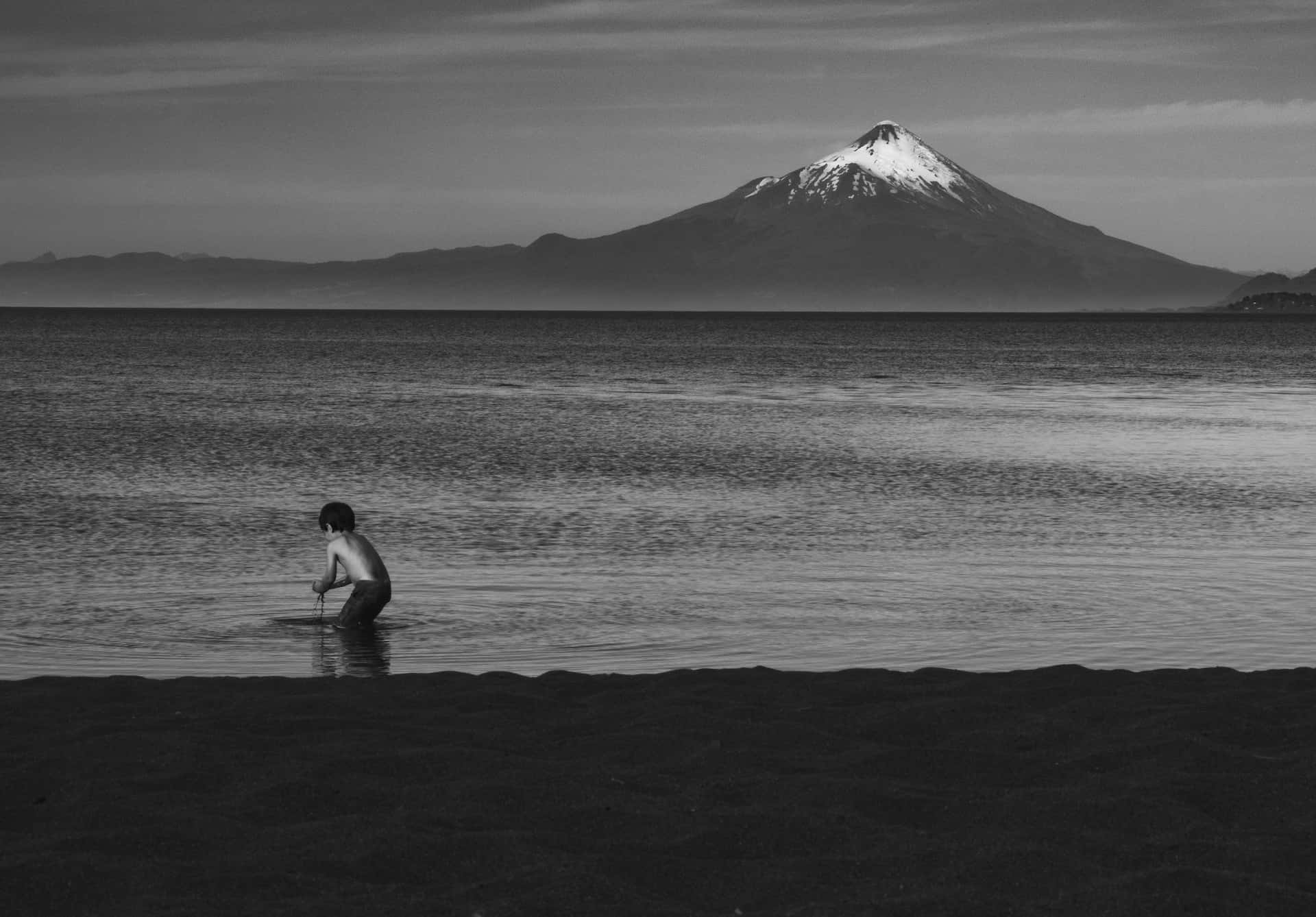 Black And White Beach 桌布 壁紙 — 一個孤獨的孩子在沙灘的淺水中涉水，遠處有一座積雪覆蓋的山峰在陰天中若隱若現。