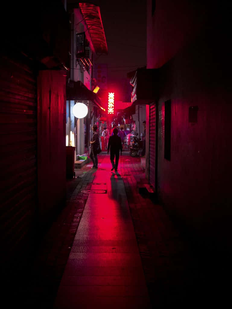 Red Light District 桌布 壁紙 — 一個孤獨的身影在夜晚走下一條狹窄的小巷，沐浴在強烈的紅色霓虹燈光中，燈光在潮濕的路面上反射。