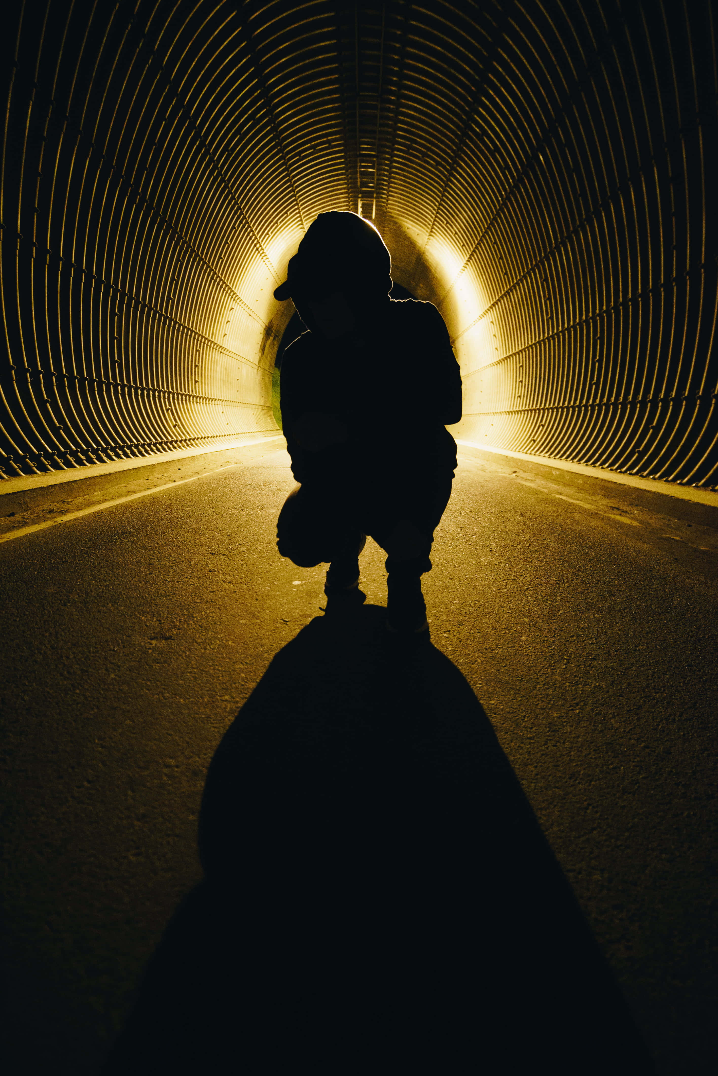 shadow 桌布 壁紙 — 一個孤獨的身影在彎曲的隧道內以剪影蹲伏，隧道被遠處出口的溫暖金色光芒照亮。