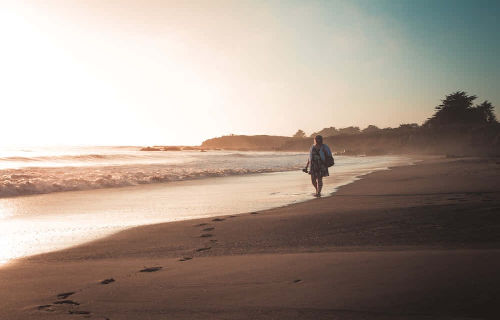 Beach Walk 桌布 壁紙 — 一個孤獨的身影在日落時分沿著陽光普照的海灘行走，在濕沙上留下腳印，海浪翻滾而來。