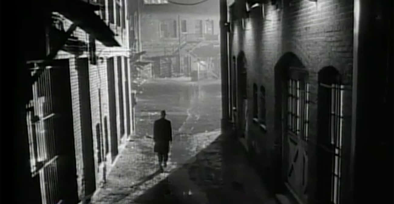 Film Noir 桌布 壁紙 — 一個孤獨的身影在昏暗的燈光下，沿著磚樓之間狹窄濕滑的巷子行走。