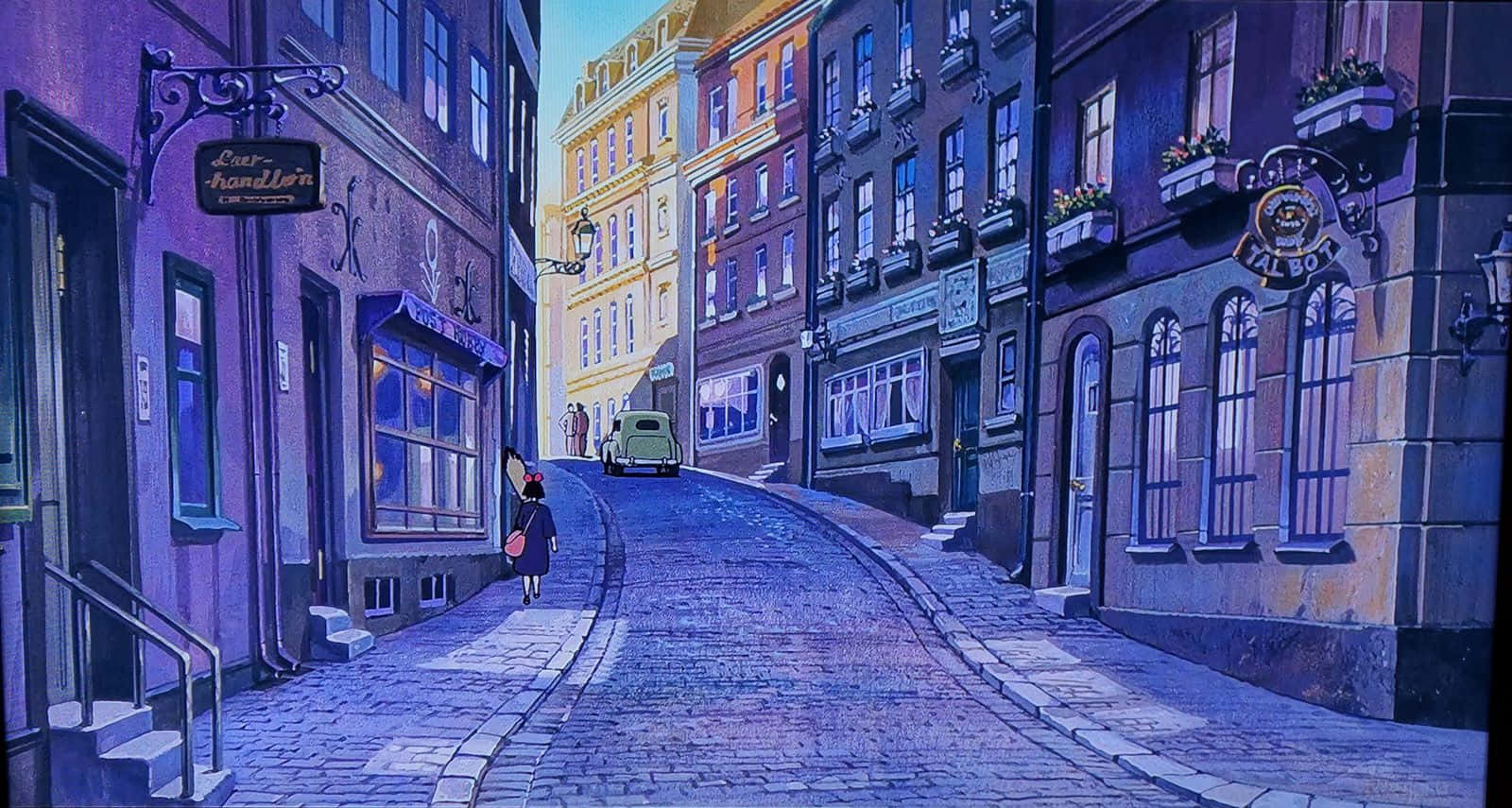kiki's delivery service 桌布 壁紙 — 一個孤獨的身影在柔和的暮色天空下，沿著一條鋪滿鵝卵石、兩旁是色彩繽紛的歐洲建築的街道行走。
