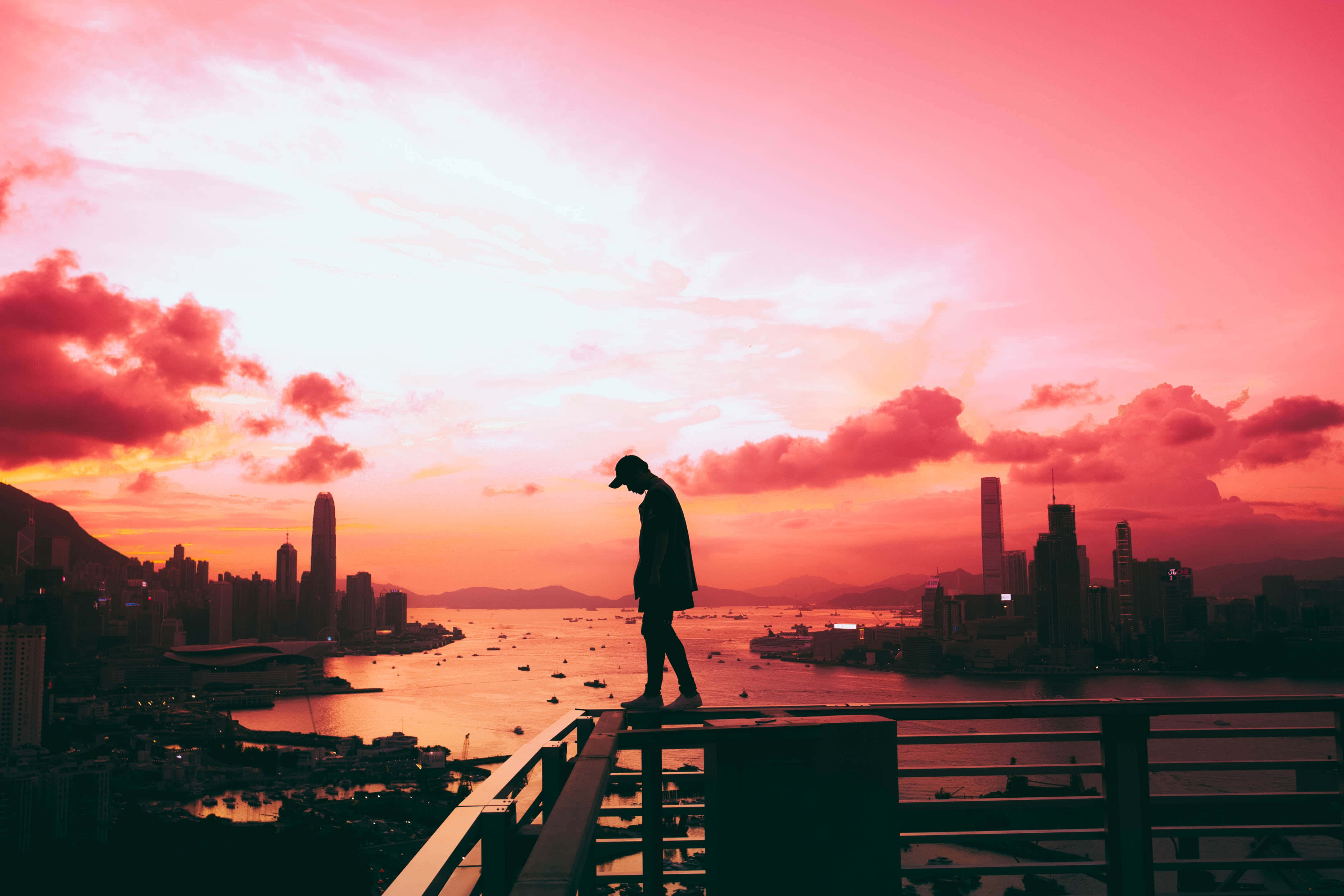 Silhouette 桌布 壁紙 — 一個孤獨的身影在欄杆上形成剪影，俯瞰著廣闊的城市景觀和海港上充滿活力的粉紅色和橙色日落。