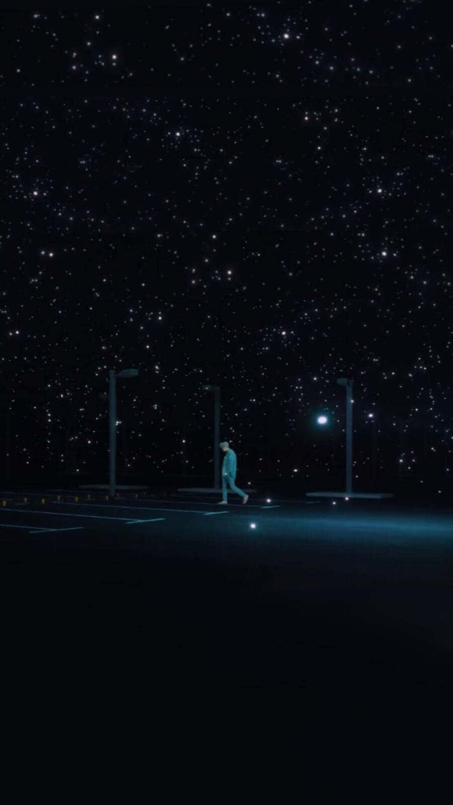 Bts Mv 桌布 壁紙 — 一個孤獨的身影在空無一人的停車場上行走，頭上是廣闊的星空，被街燈照亮。