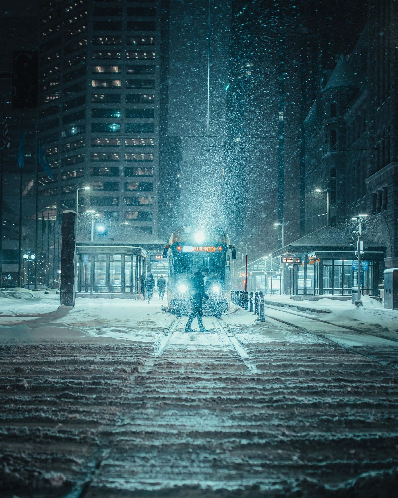 Snowstorm 桌布 壁紙 — 一個孤獨的身影在雪覆蓋的夜晚城市街道上，走向一輛發光的巴士，周圍是高聳的建築物和飄落的雪花。