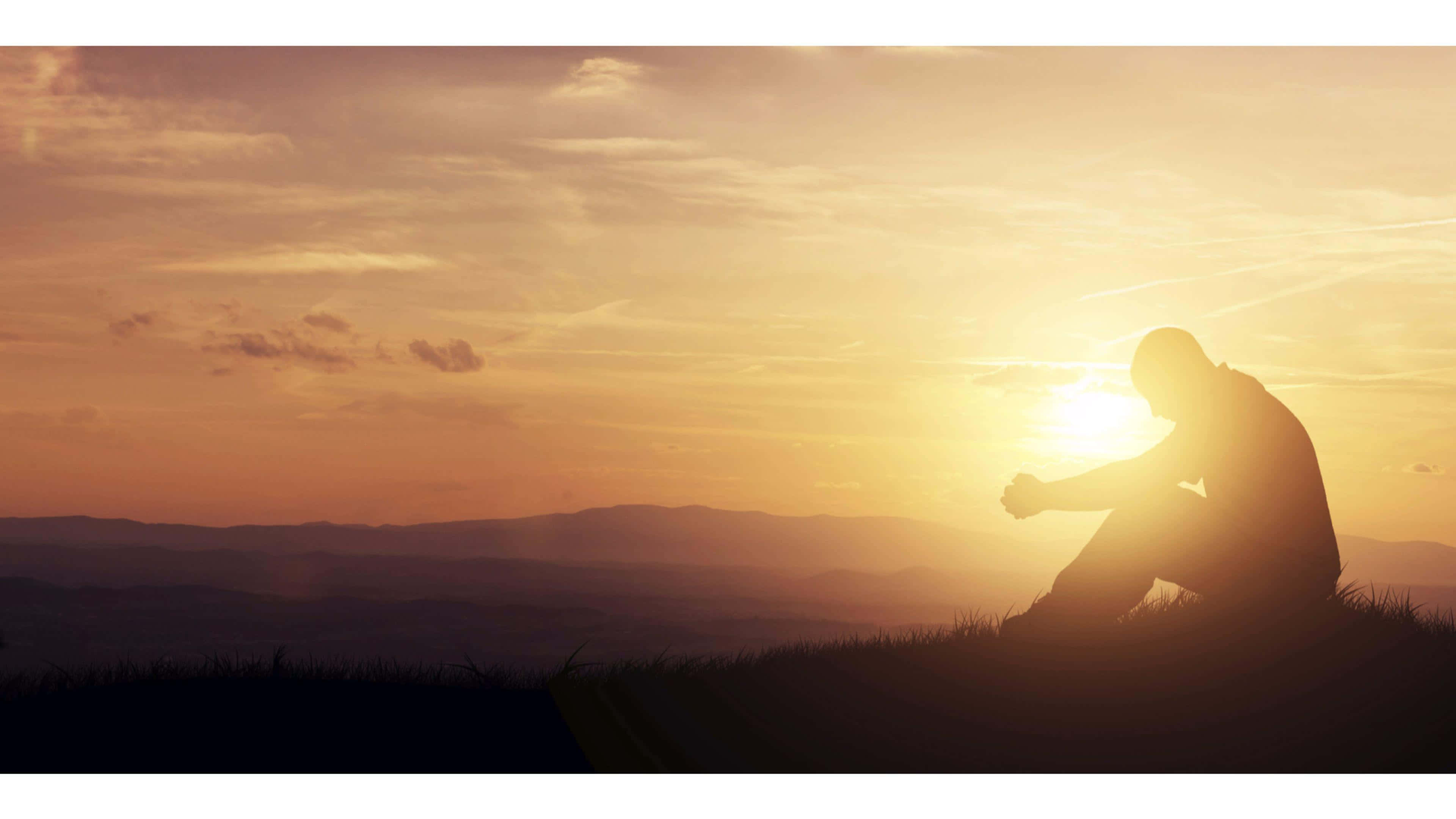 Praying 桌布 壁紙 — 一個孤獨的身影坐在山頂上沉思，夕陽在他們身後落下，將溫暖的金色光芒投射在遠山之上。