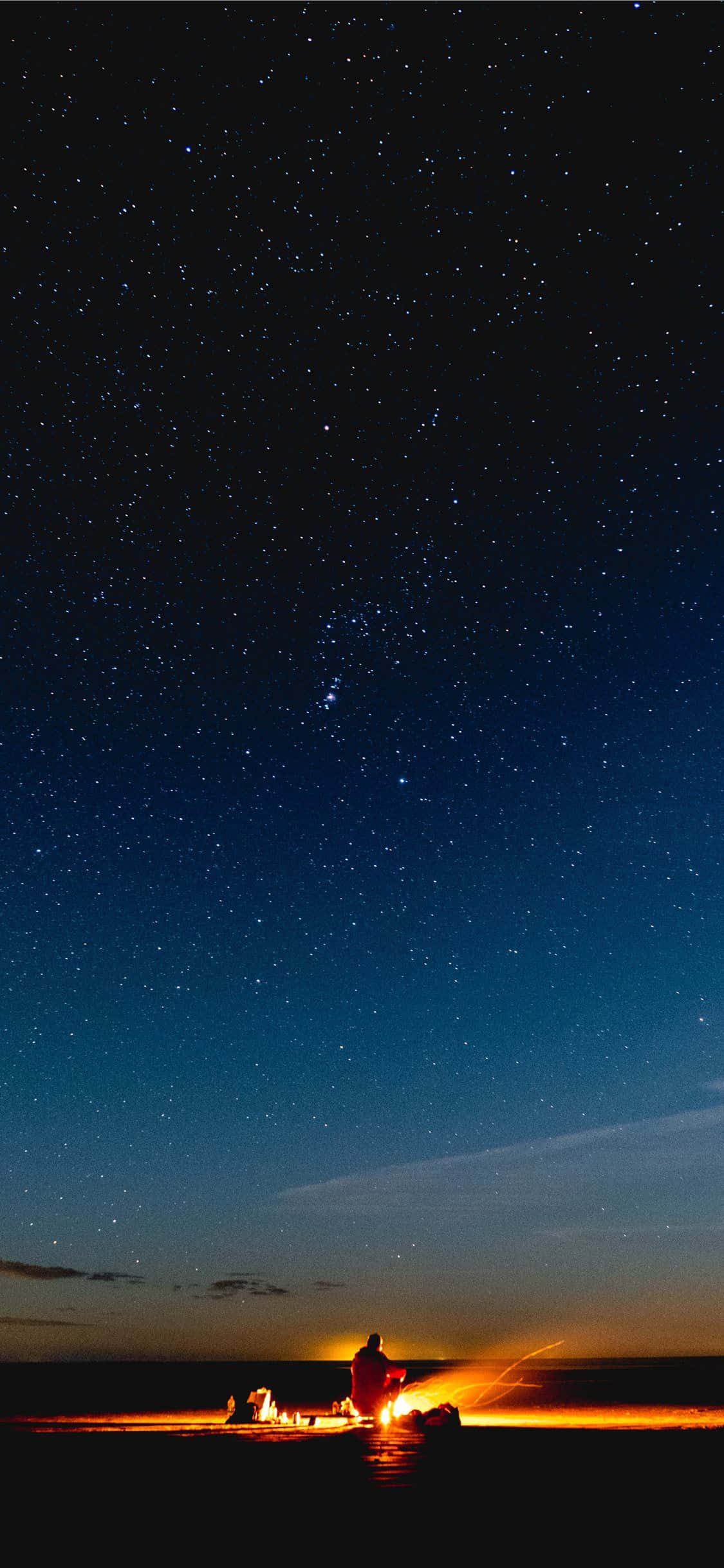 Beach Bonfire 桌布 壁紙 — 一個孤獨的身影坐在黑暗海灘上的營火旁，上方是廣闊的星空。