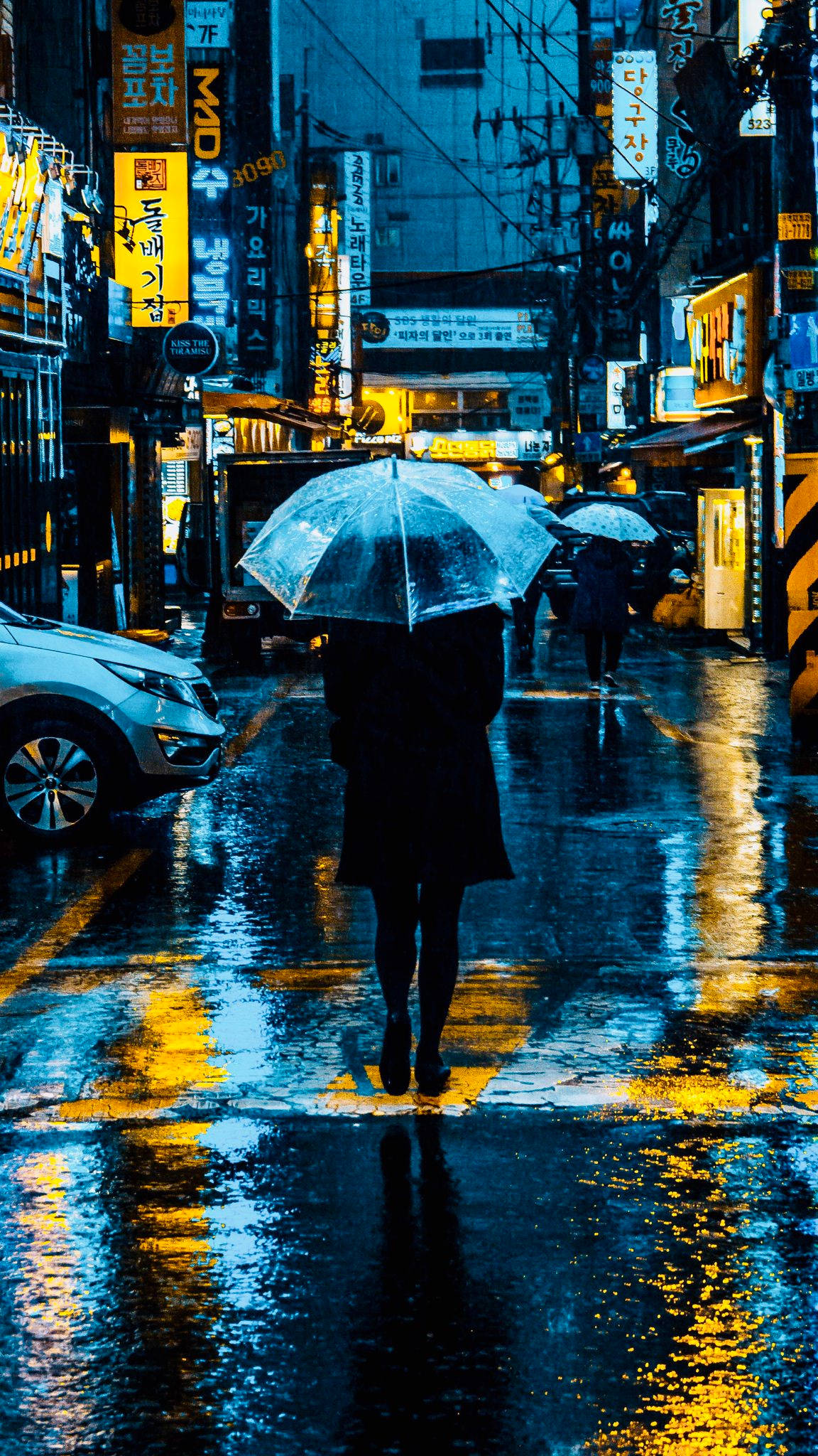 東京 桌布 壁紙 — 一個孤獨的身影撐著傘，走過一條被雨水浸濕、霓虹燈閃爍的韓國街道，反射出鮮黃色和深藍色的燈光。