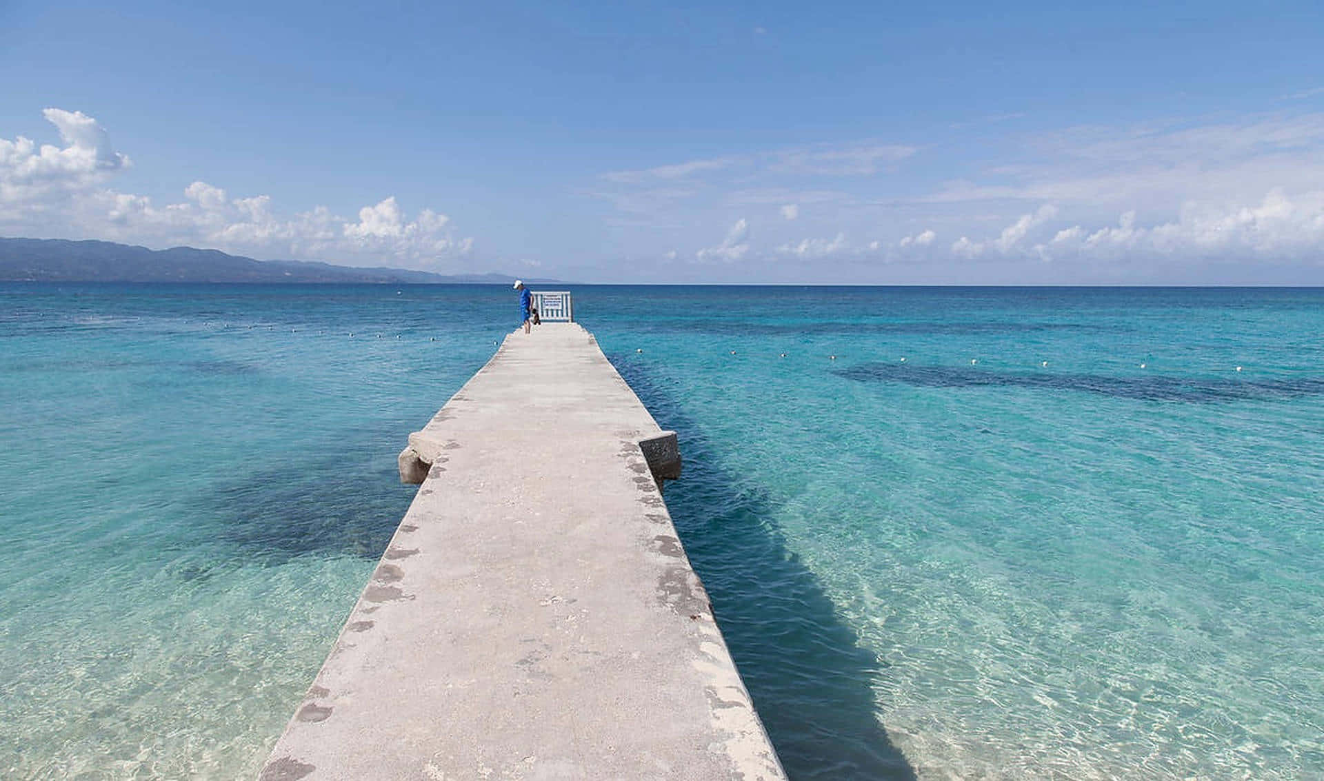 Jamaica 桌布 壁紙 — 一個孤獨的身影沿著一條長長的混凝土碼頭行走，碼頭延伸至清澈碧綠的海洋，上方是帶有零星白雲的明亮藍天。