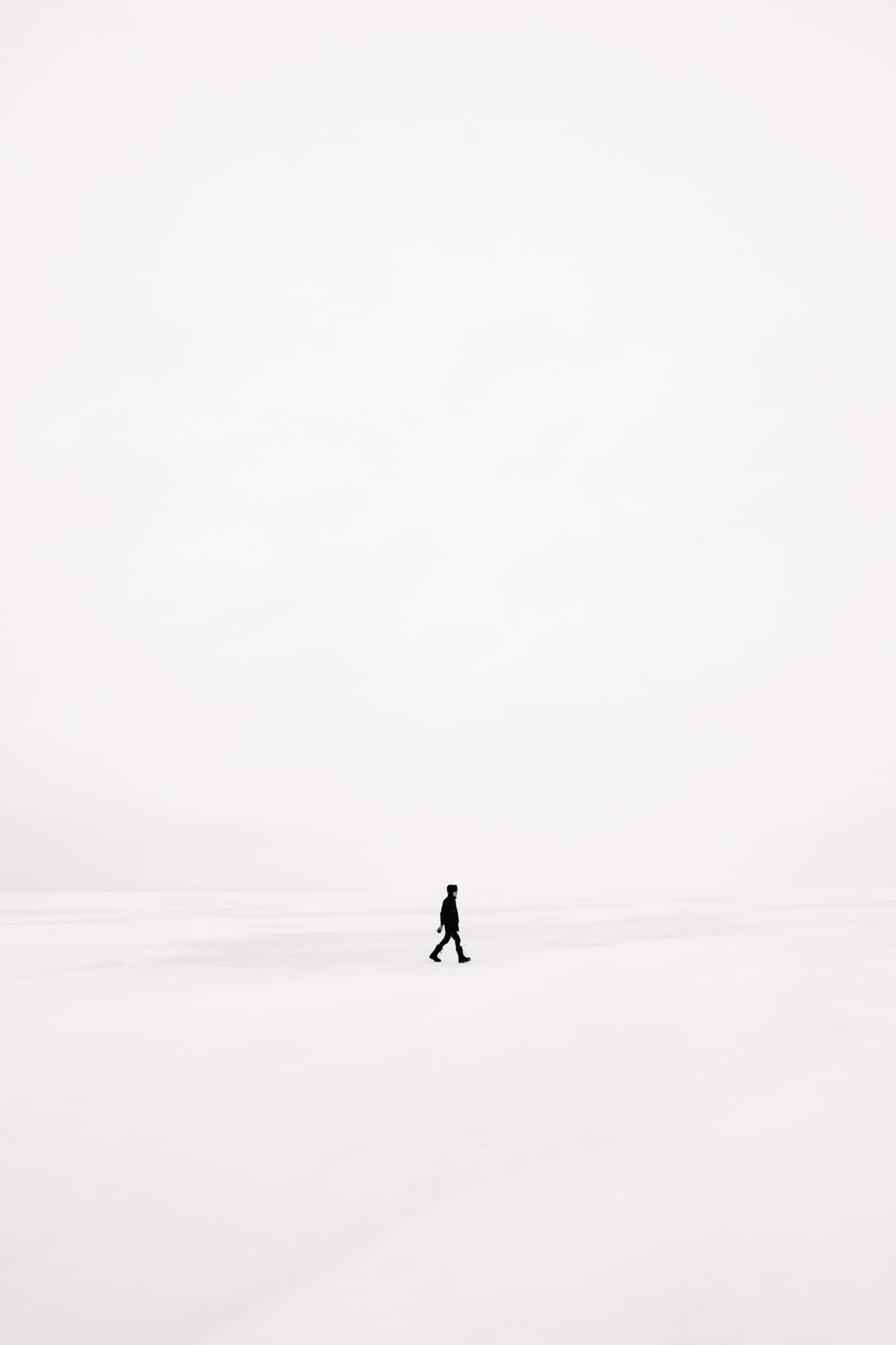 白色 iPhone 桌布 壁紙 — 一個孤獨的身影獨自走過無盡的雪原，天空蒼白多雲。