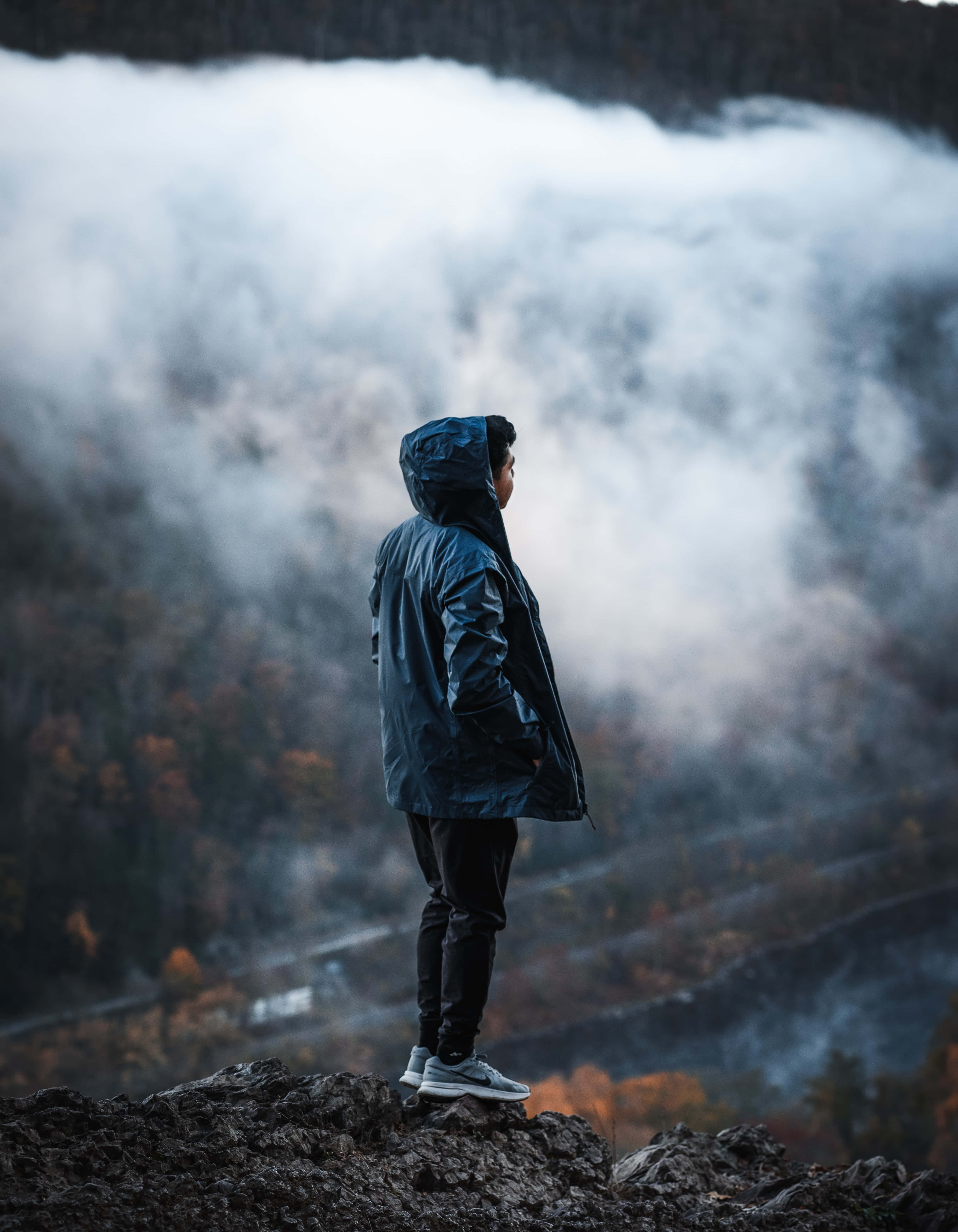 Navy Blue Jacket 桌布 壁紙 — 一個孤獨的身影穿著連帽外套，站在岩石峭壁上，俯瞰著被薄霧籠罩的山谷，下方有秋天的樹木。