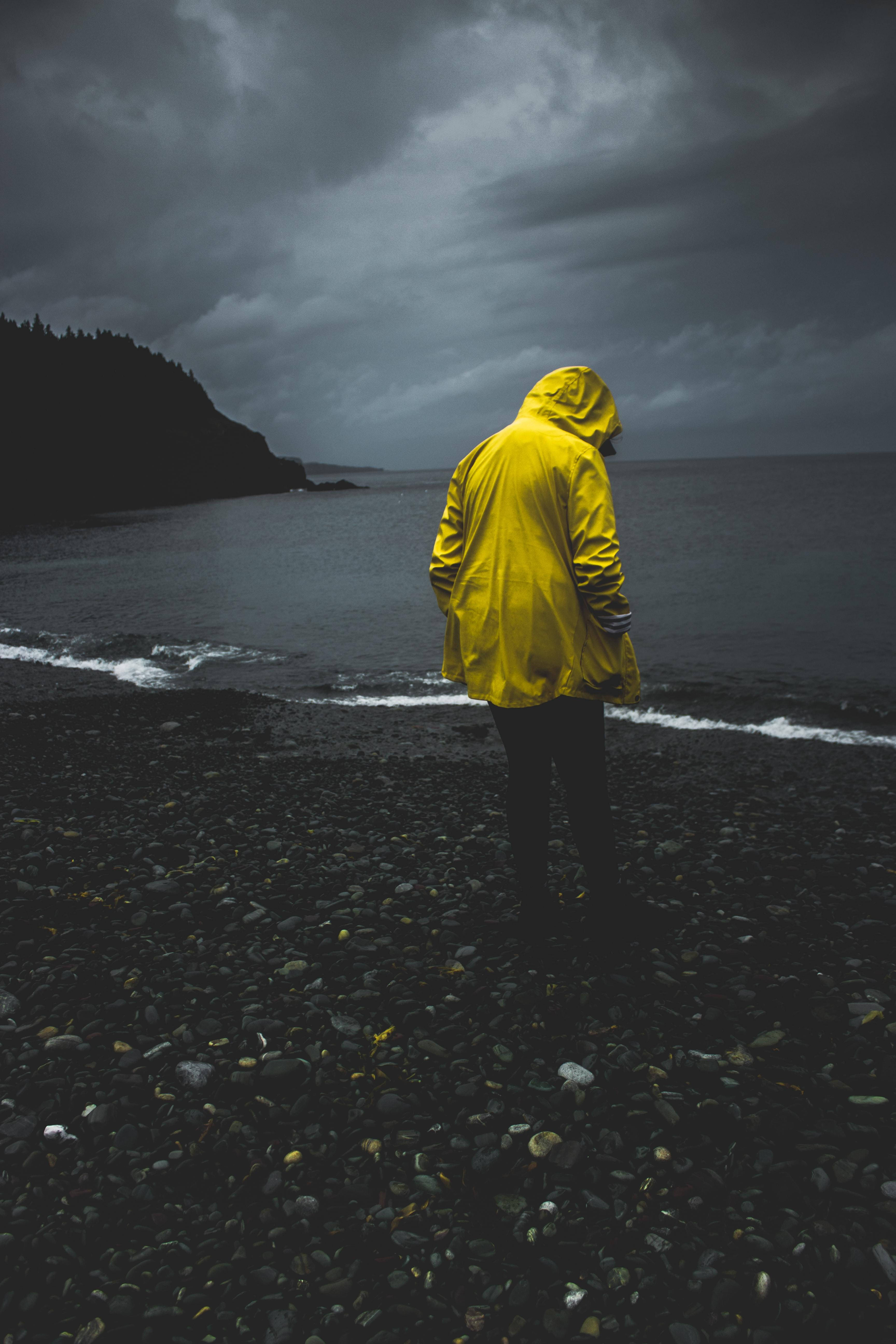 海灘 桌布 壁紙 — 一個孤獨的身影穿著鮮黃色雨衣，站在黑暗的卵石海灘上，面對著厚重烏雲下的暴風雨大海。