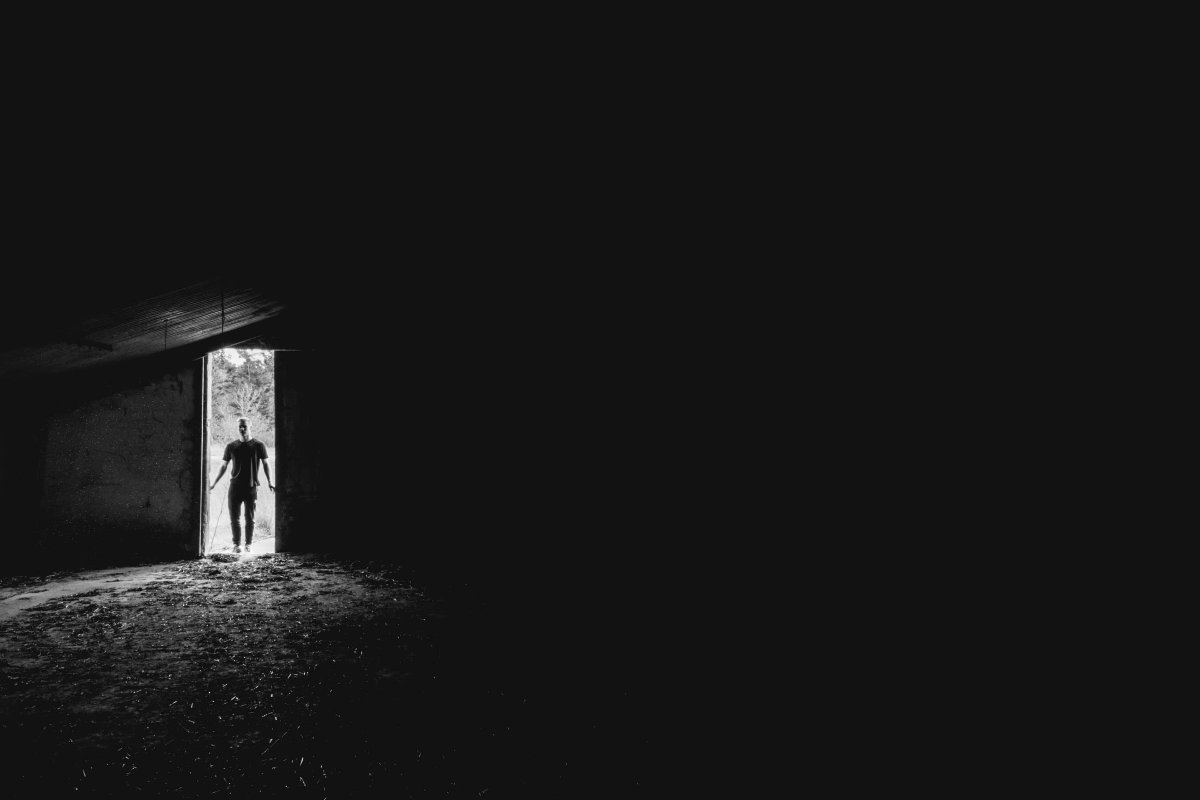 Dark Room 桌布 壁紙 — 一個孤獨的身影站在門口，沐浴在明亮的光線中，在黑暗空蕩的室內襯托下形成剪影。