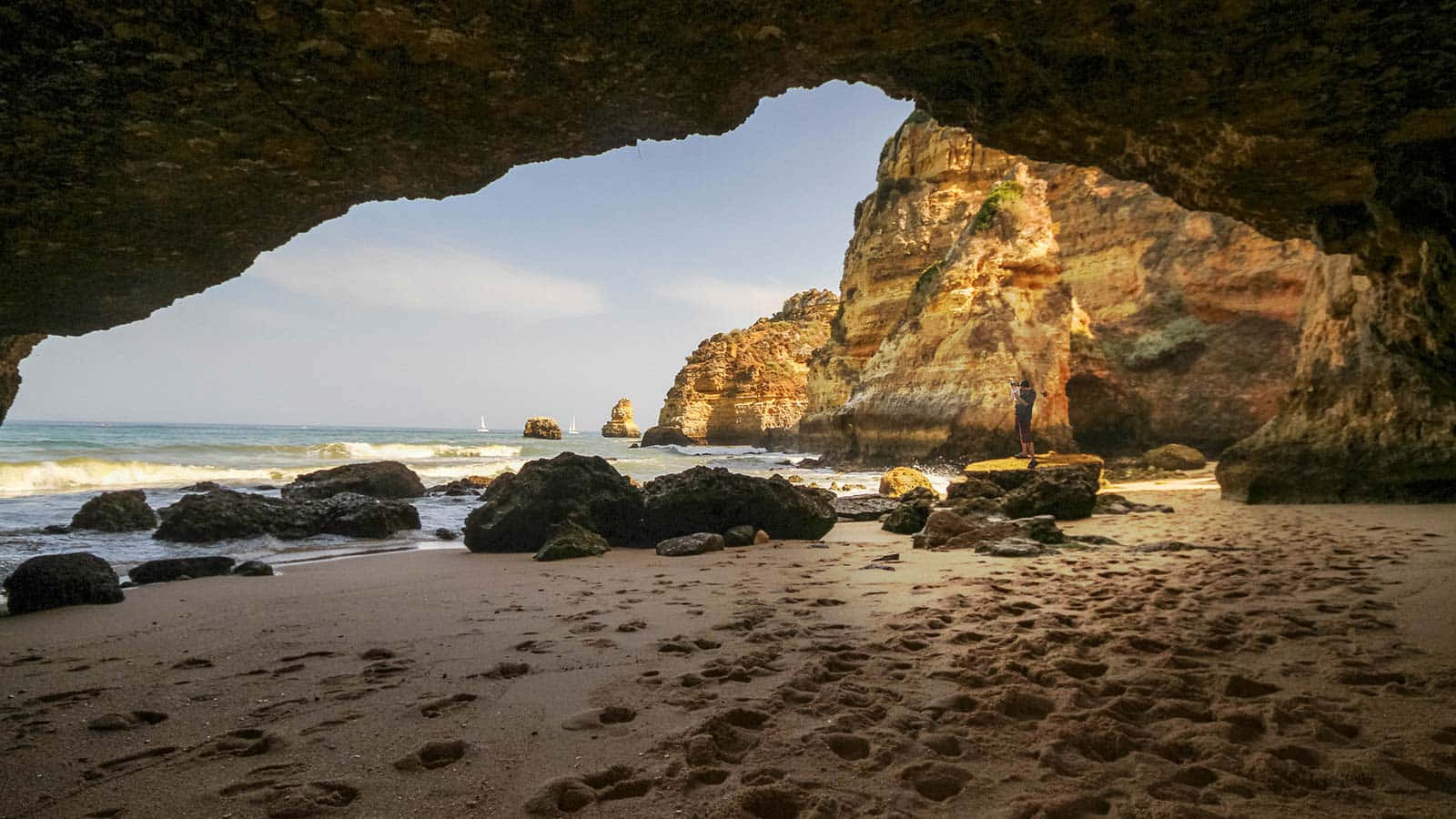 Portuguese Beach 桌布 壁紙 — 一個孤獨的身影站在陽光明媚的沙灘上，被天然岩石拱門框住，俯瞰著碧綠的海浪和崎嶇的懸崖。