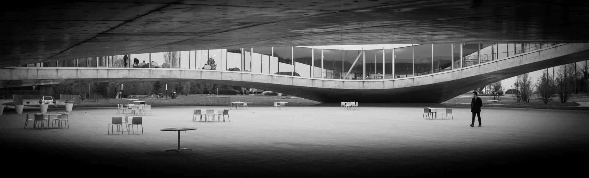 Black And White Architecture 桌布 壁紙 — 一個孤獨的身影行走在巨大的彎曲混凝土結構下，廣場上散落著椅子和桌子，呈現出 stark 的黑白城市廣場景象。