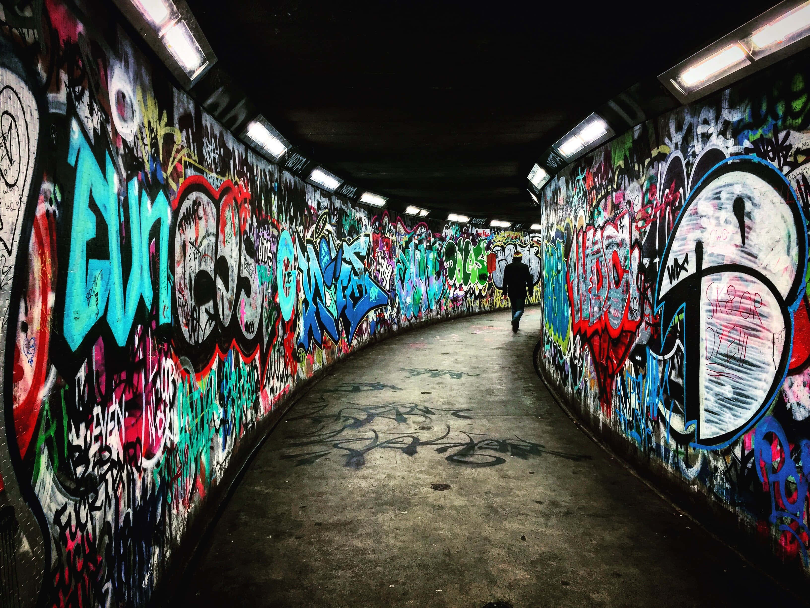 Dark Underground 桌布 壁紙 — 一個孤獨的身影走過一條彎曲的隧道，隧道內壁在螢光燈下佈滿了鮮豔的塗鴉。
