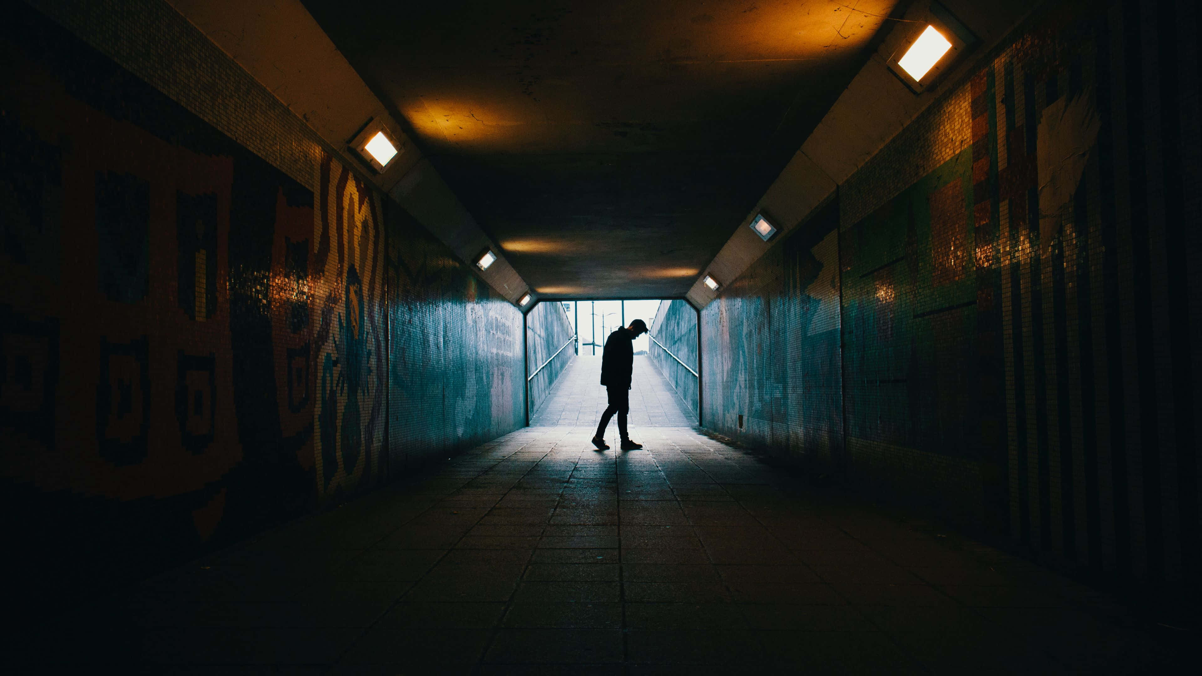 Dark Underground 桌布 壁紙 — 一個孤獨的身影走過一條燈光昏暗的城市隧道，牆上有塗鴉，頭頂有溫暖的燈光。