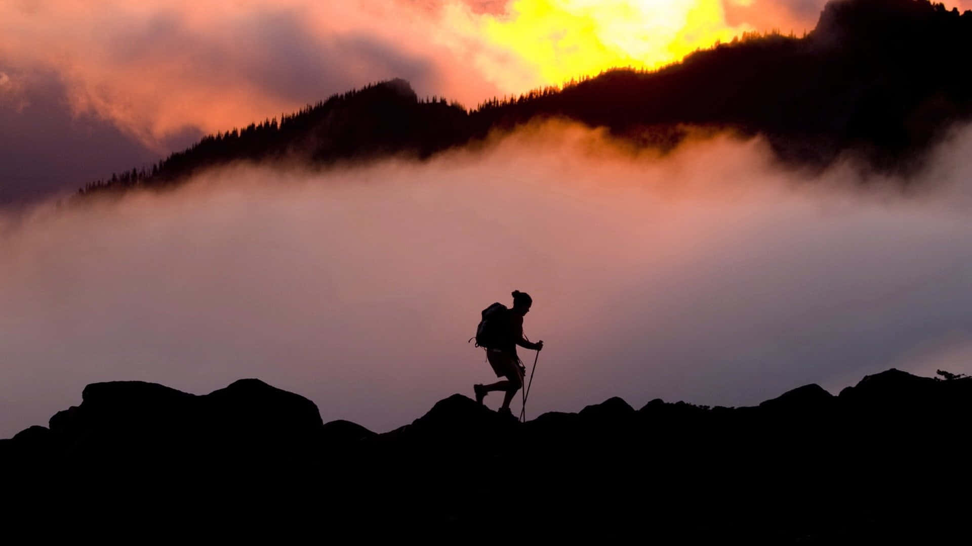 Hiking 桌布 壁紙 — 一個孤獨的遠足者的剪影攀爬岩石山峰，熾熱的日落光線以溫暖的色調描繪雲彩和遠處的山脈。