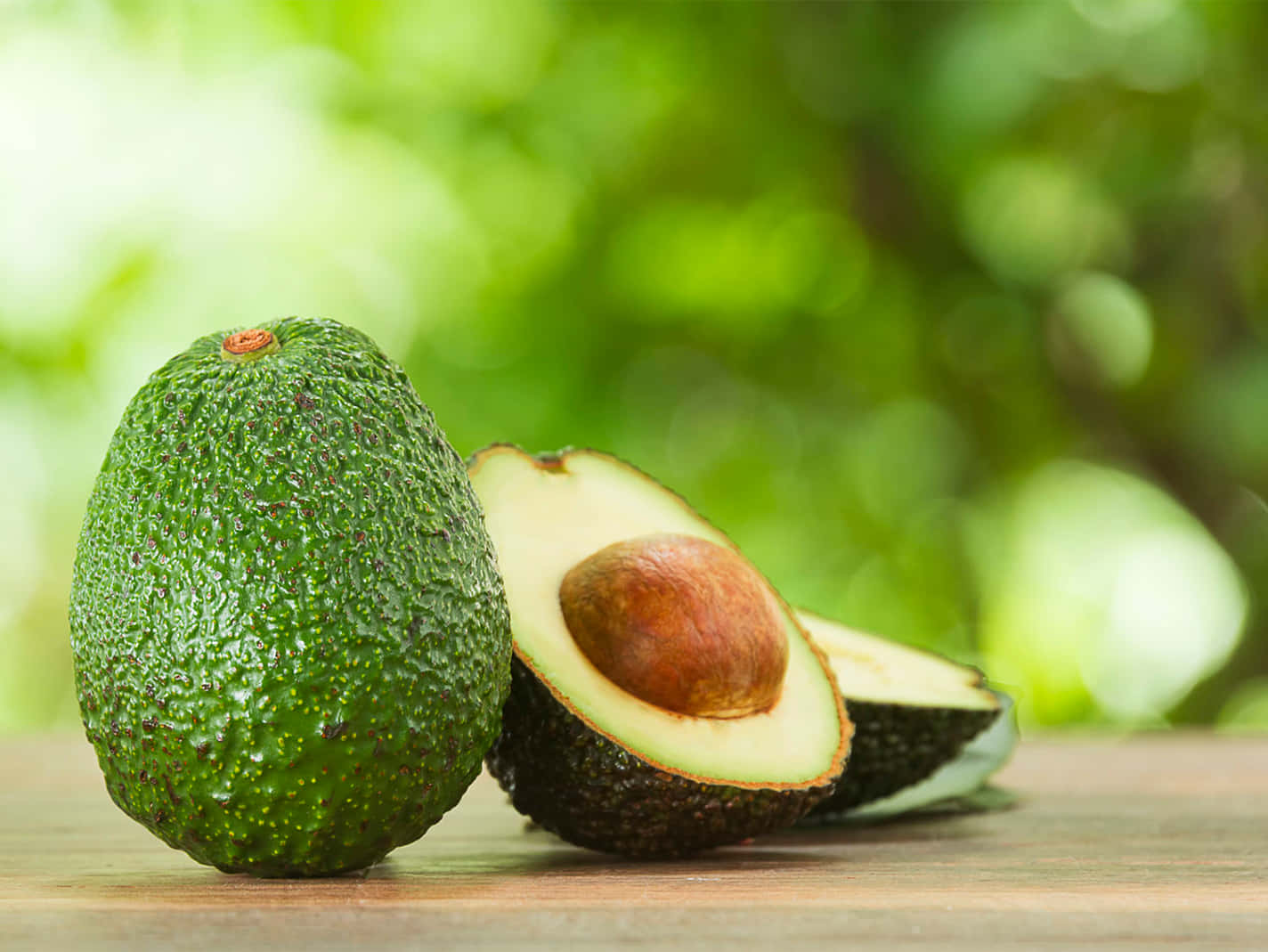 Avocado 桌布 壁紙 — 一個完整和切半的牛油果，帶有棕色果核，放在木質表面上，背景是柔和的綠色模糊效果。
