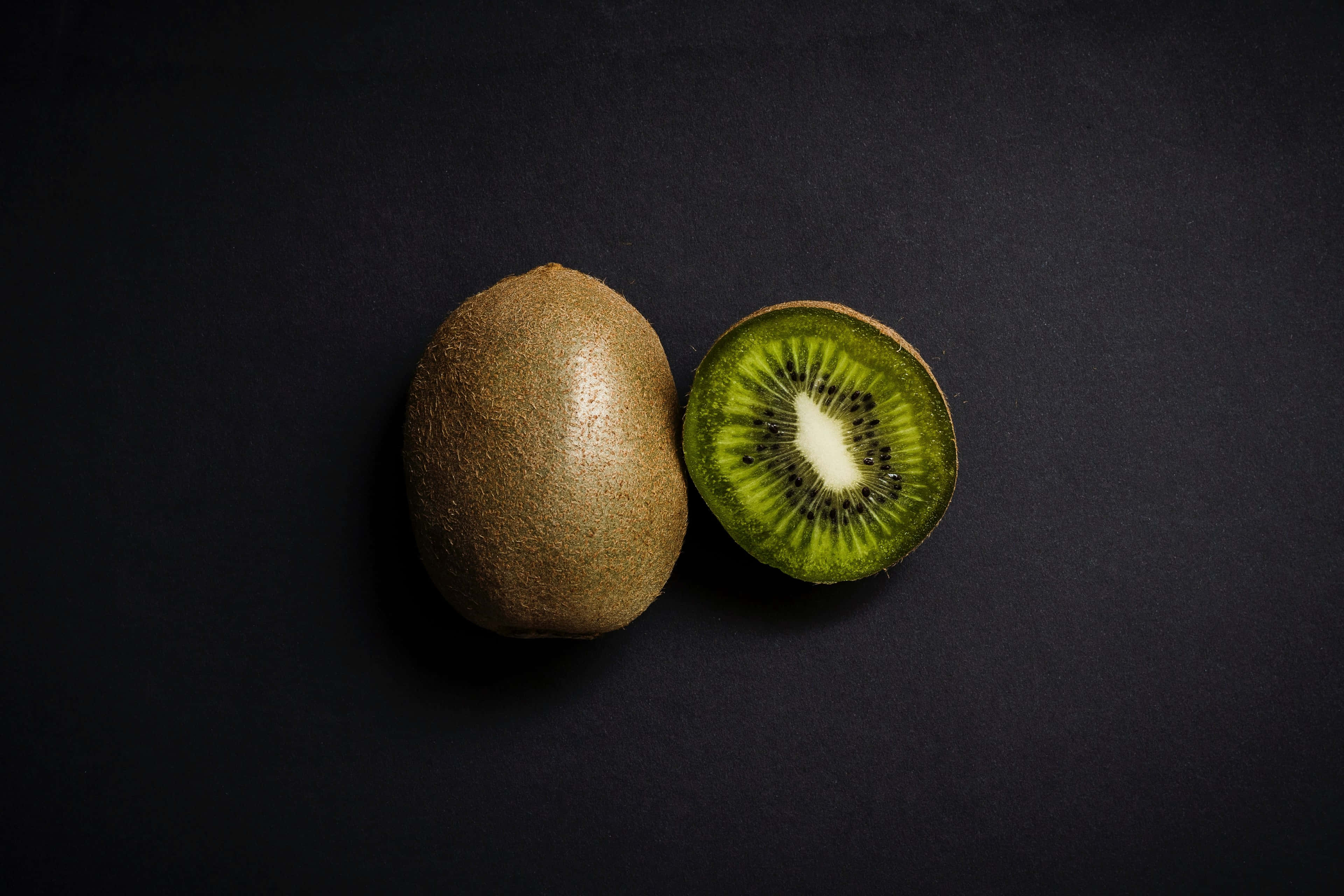 Kiwi 桌布 壁紙 — 一個完整的奇異果和一個切片的奇異果，在深色紋理背景上展現出鮮綠色的果肉和黑色的種子。