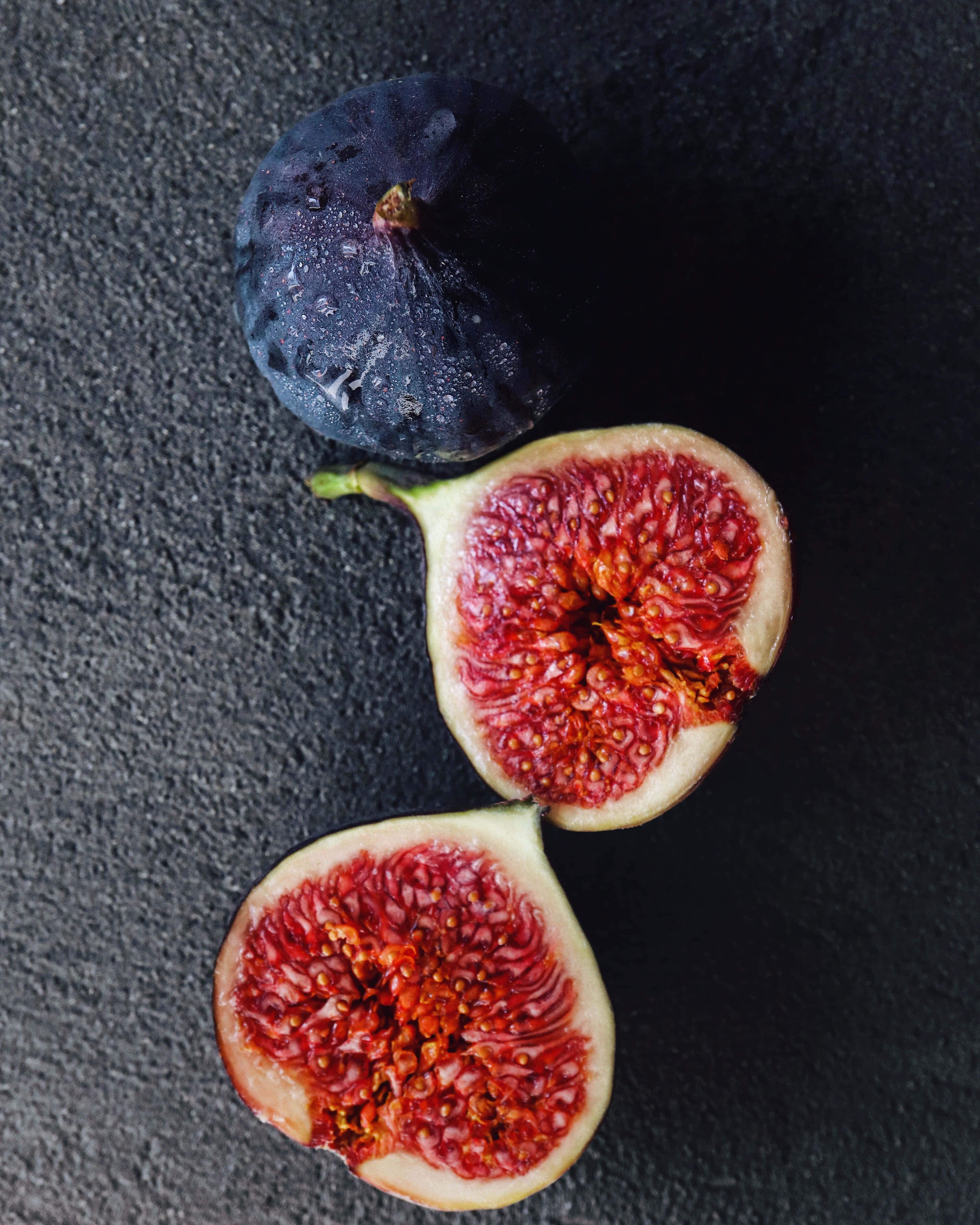 Purple Figs 桌布 壁紙 — 一個完整的深色無花果和兩個切半的無花果，露出多汁的紅色果肉，放在有紋理的黑色表面上。