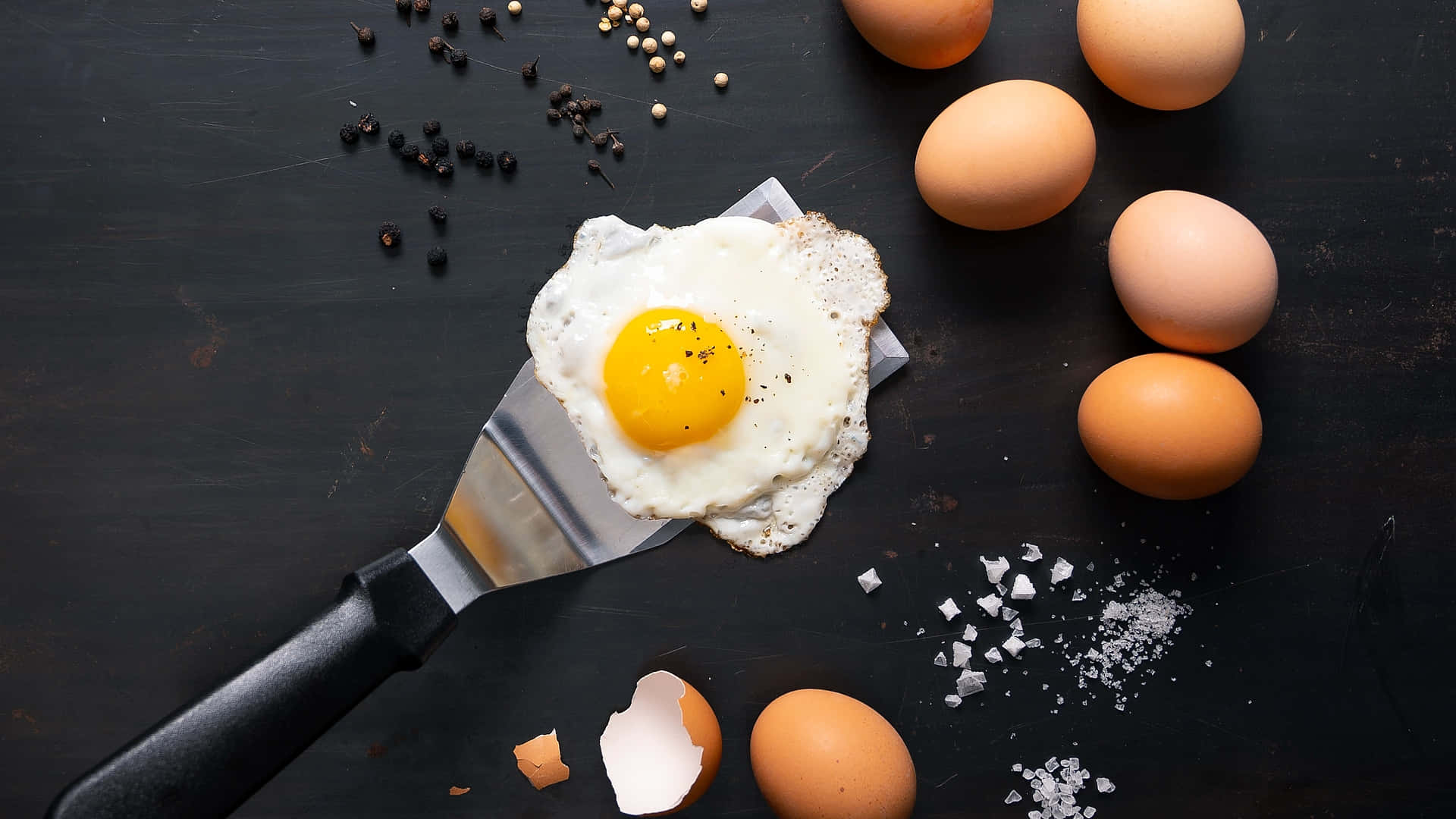 Egg White 桌布 壁紙 — 一個完美煎好的太陽蛋放在鑊鏟上，周圍有全蛋、鹽和胡椒，置於深色鄉村風格的表面上。