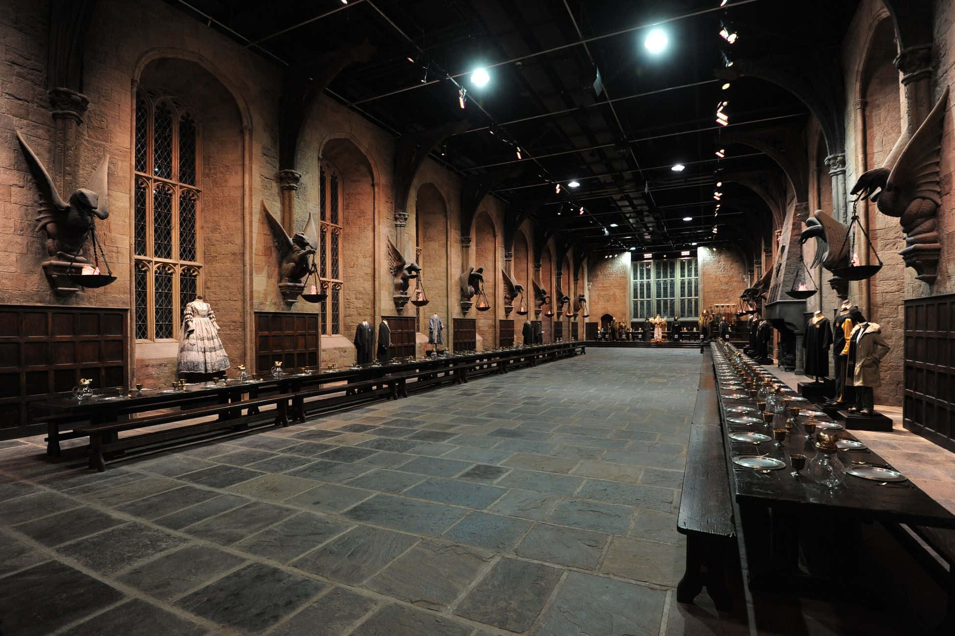 The Hogwarts Great Hall 桌布 壁紙 — 一個宏偉、燈光昏暗的中世紀宴會廳，舖有石地板、拱形窗戶，並在裸露的橫樑和聚光燈下擺放著長桌，準備舉行盛宴。