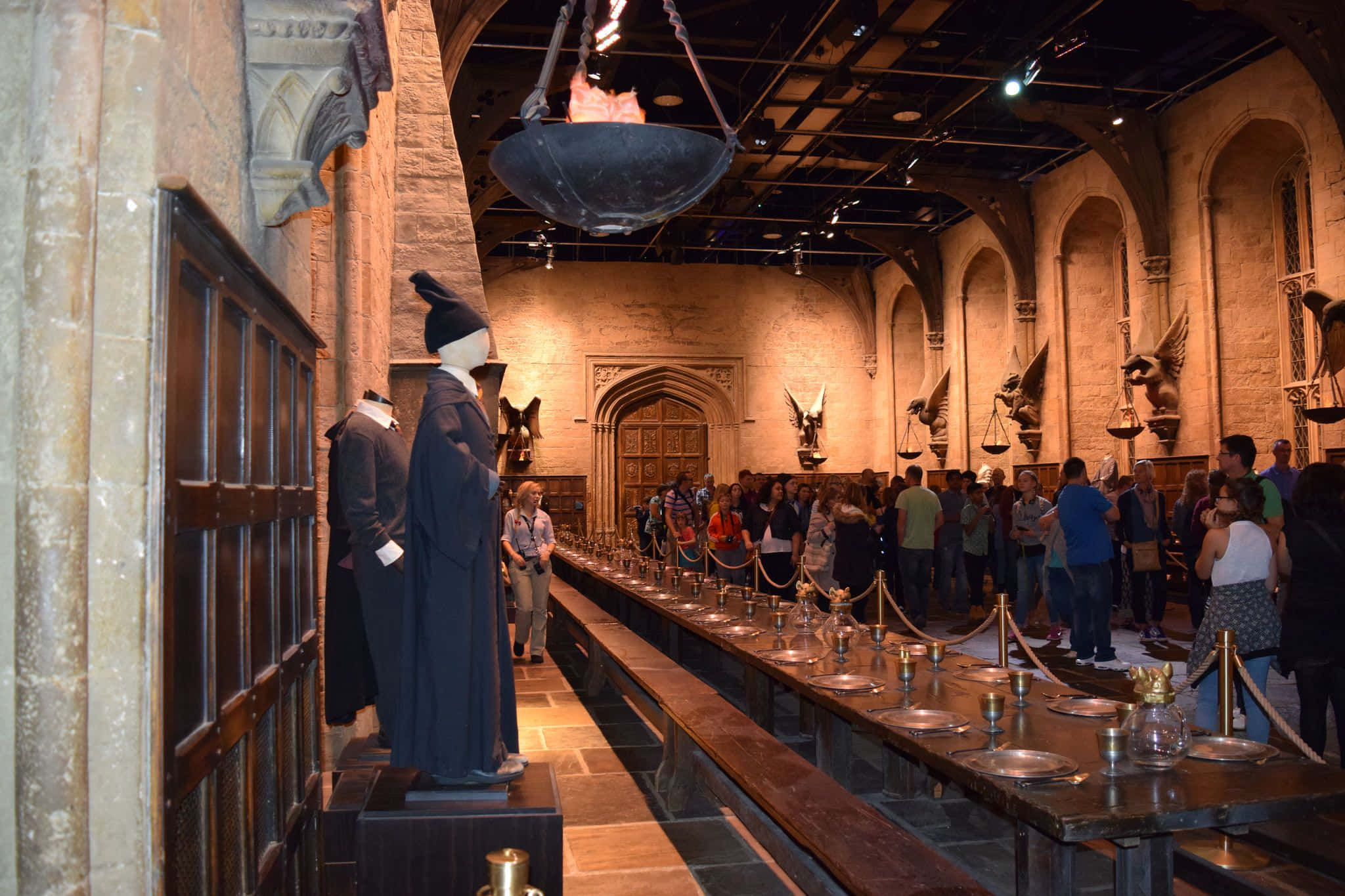 The Hogwarts Great Hall 桌布 壁紙 — 一個宏偉的中世紀風格餐廳，有長木桌、掛著的鍋，以及探索主題展覽的遊客。