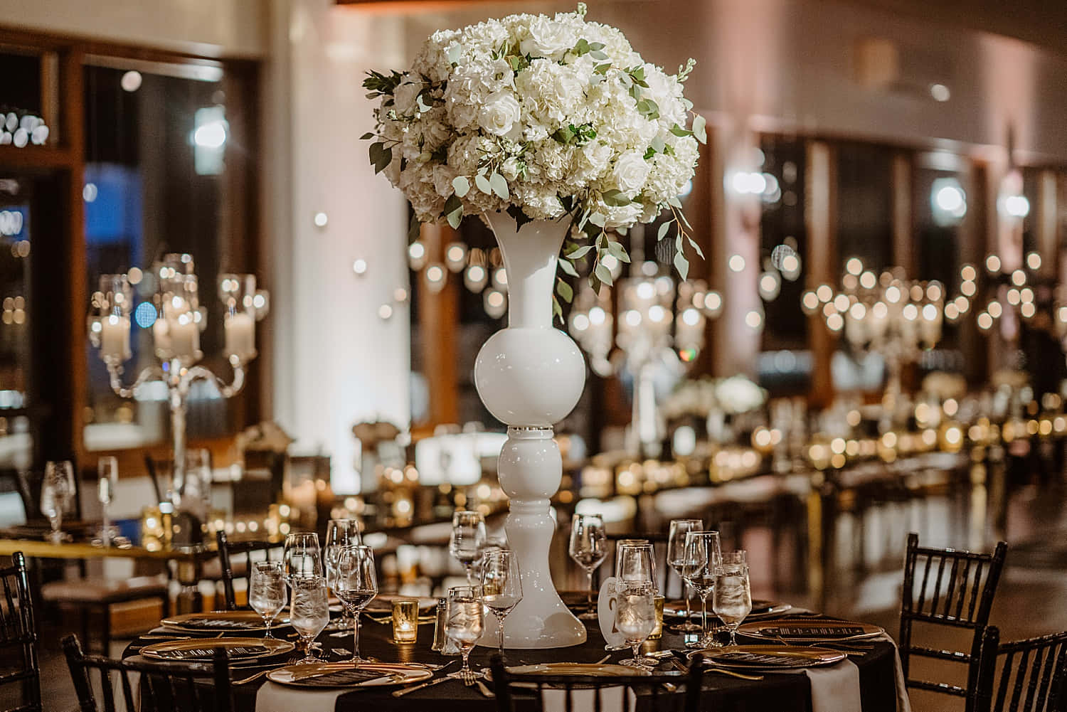 Black And White Wedding 桌布 壁紙 — 一個宏偉的宴會廳，擺放著優雅的餐桌和一個在溫暖環境燈光下閃耀的高聳白色花卉中心裝飾。