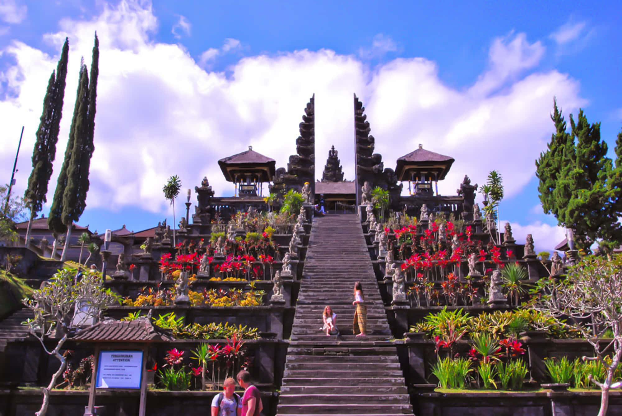 Bali 桌布 壁紙 — 一個宏偉的峇里島寺廟建築群，擁有華麗的石階，兩旁是鮮豔的紅色花朵和茂密的綠色植物，在明亮的藍天下。