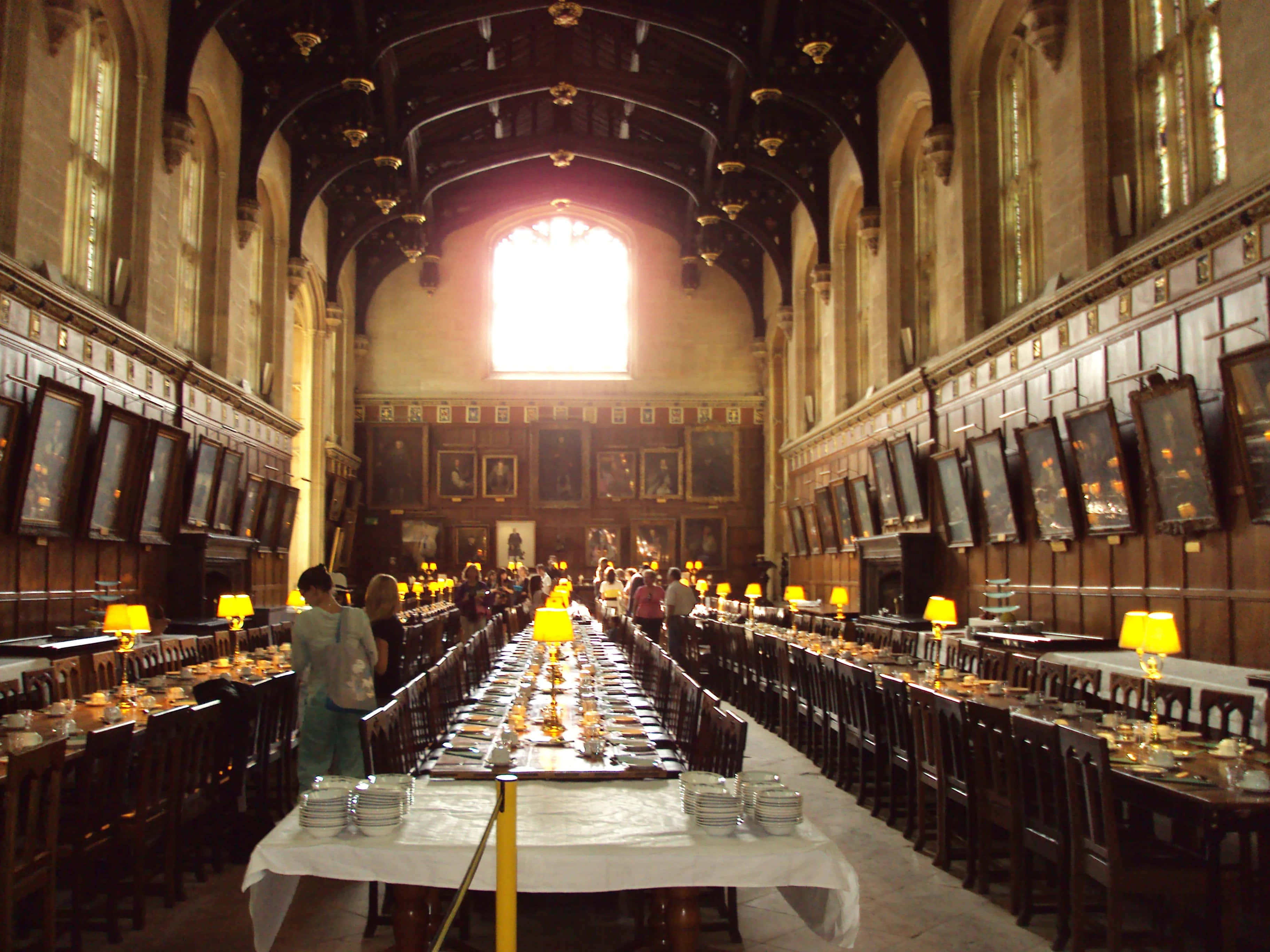 The Hogwarts Great Hall 桌布 壁紙 — 一個宏偉的歷史宴會廳，長木桌擺好餐具準備用餐，溫暖的黃色燈光亮起，陽光透過大型拱形窗戶灑進來。