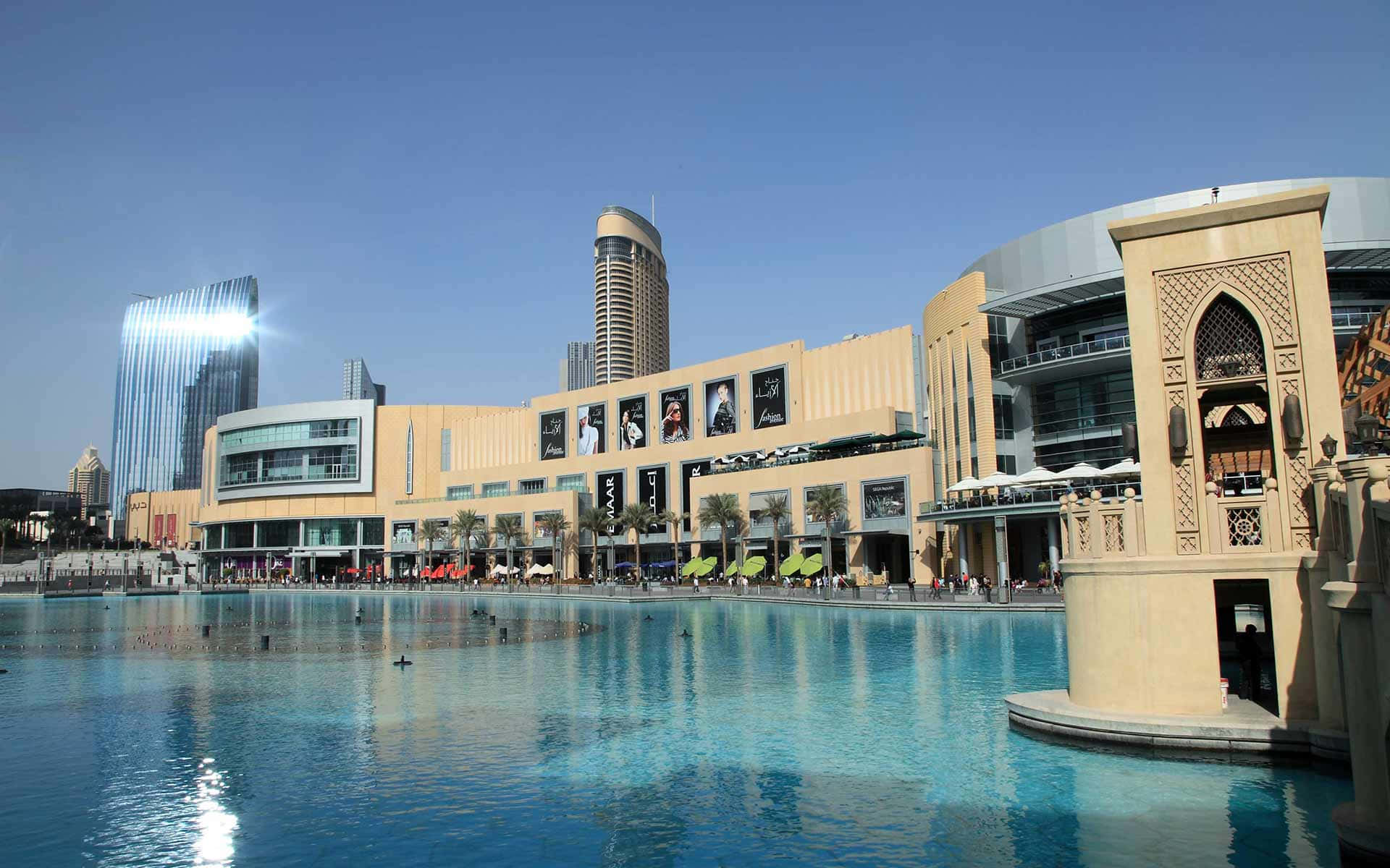 Shopping Mall 桌布 壁紙 — 一個宏偉的現代購物中心，帶有阿拉伯建築細節，在晴朗的藍天下倒映在一個大的藍綠色水池中。