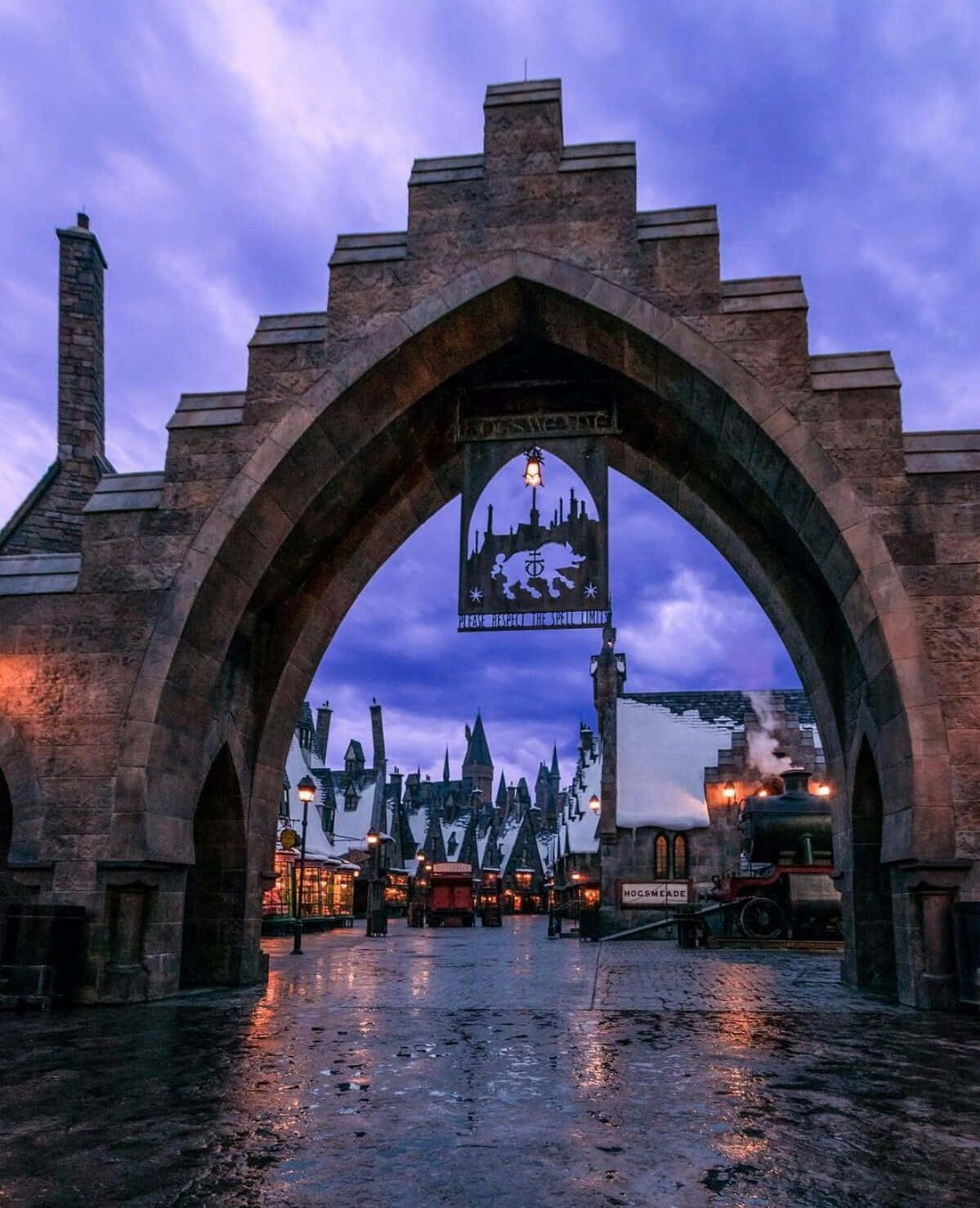 Hogsmeade 桌布 壁紙 — 一個宏偉的石拱門框住了一個黃昏時分、有蒸汽火車和發光燈籠的雪中巫師村莊。