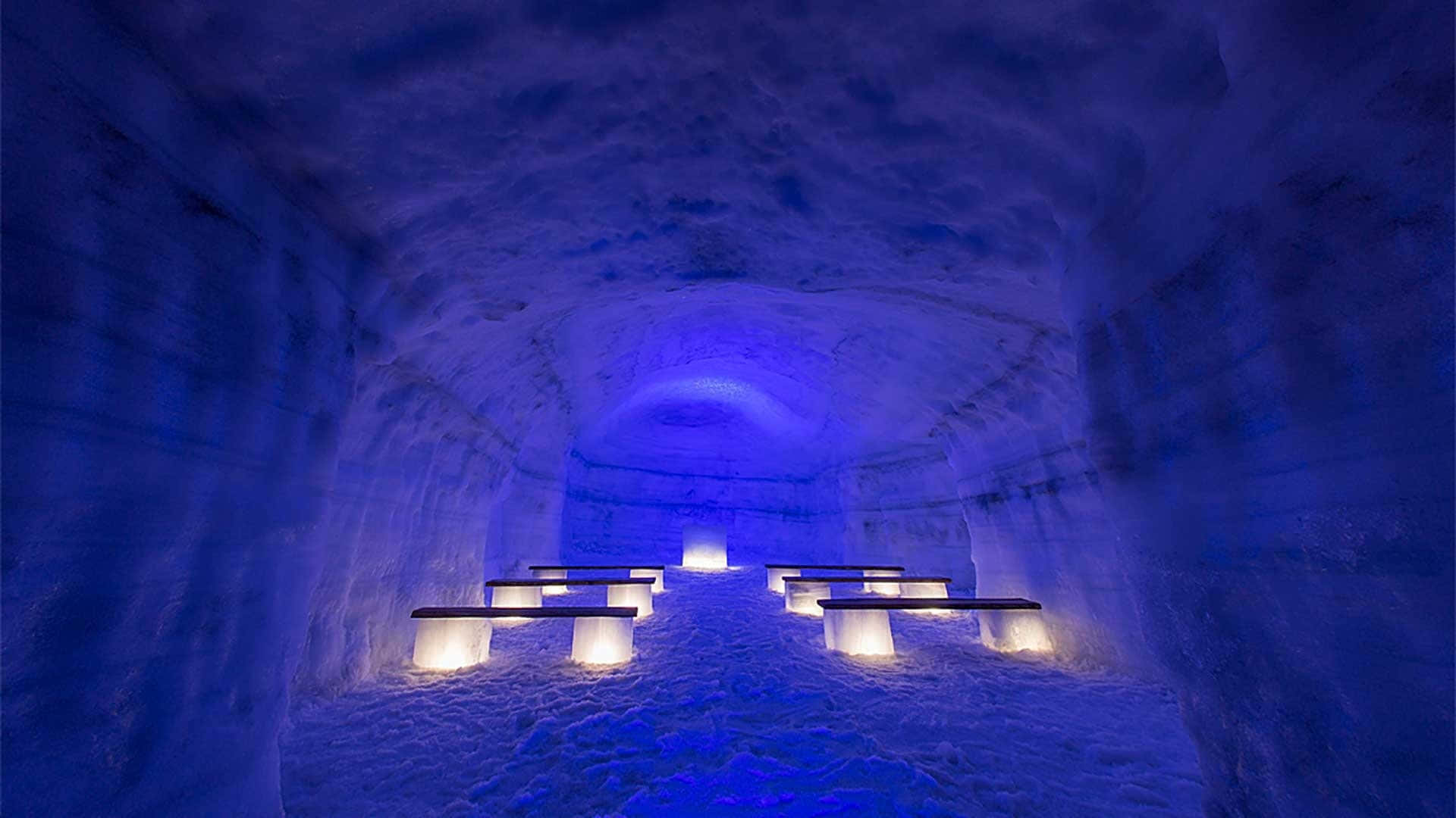 Ice Hotel 桌布 壁紙 — 一個寧靜的冰洞，發光的長椅沐浴在清涼的藍光中，營造出一個平靜的冰凍聖所。