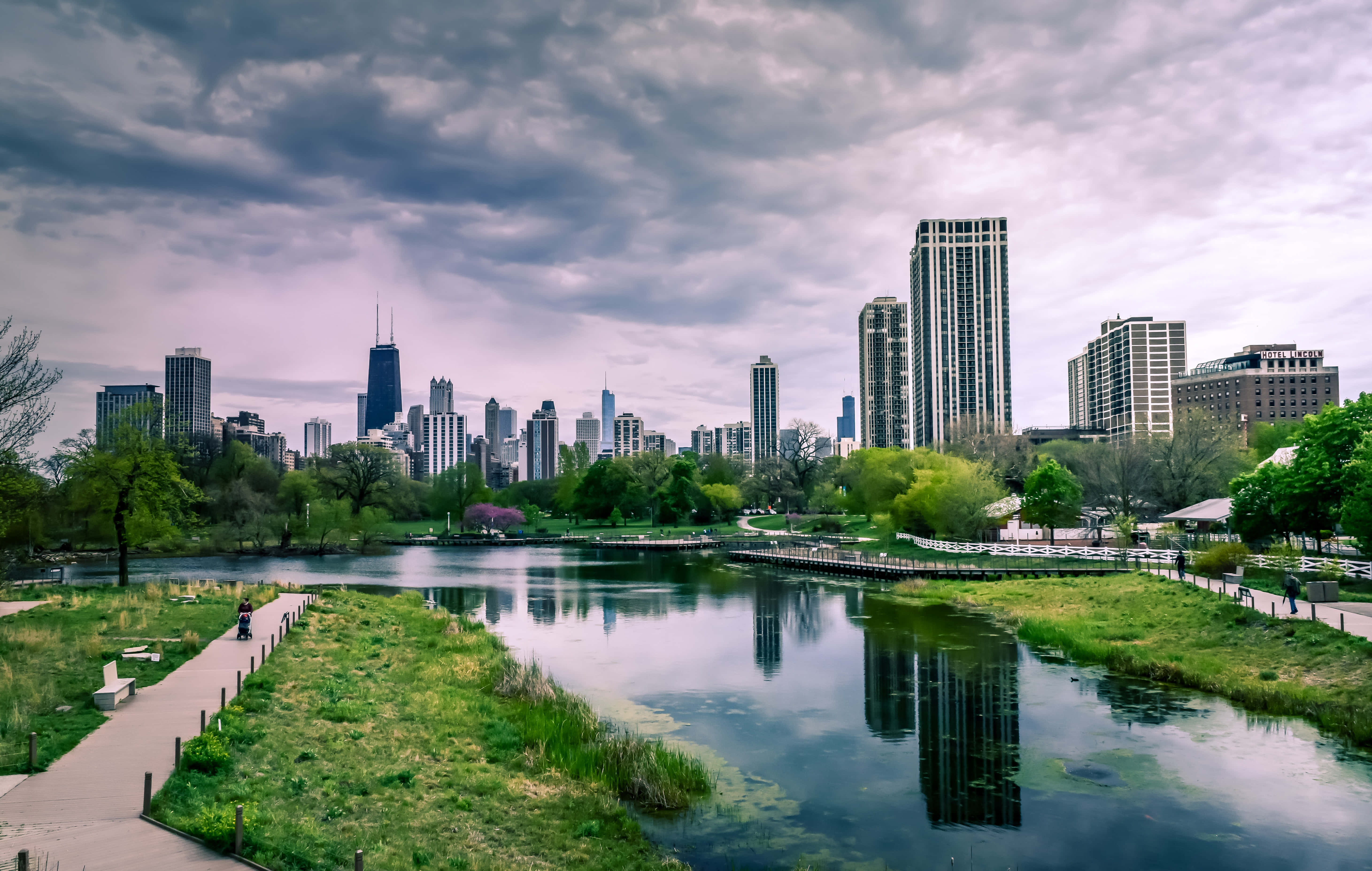 Cityscape 桌布 壁紙 — 一個寧靜的城市公園，平靜的湖面倒映著陰沉多雲天空下的高聳摩天大樓。
