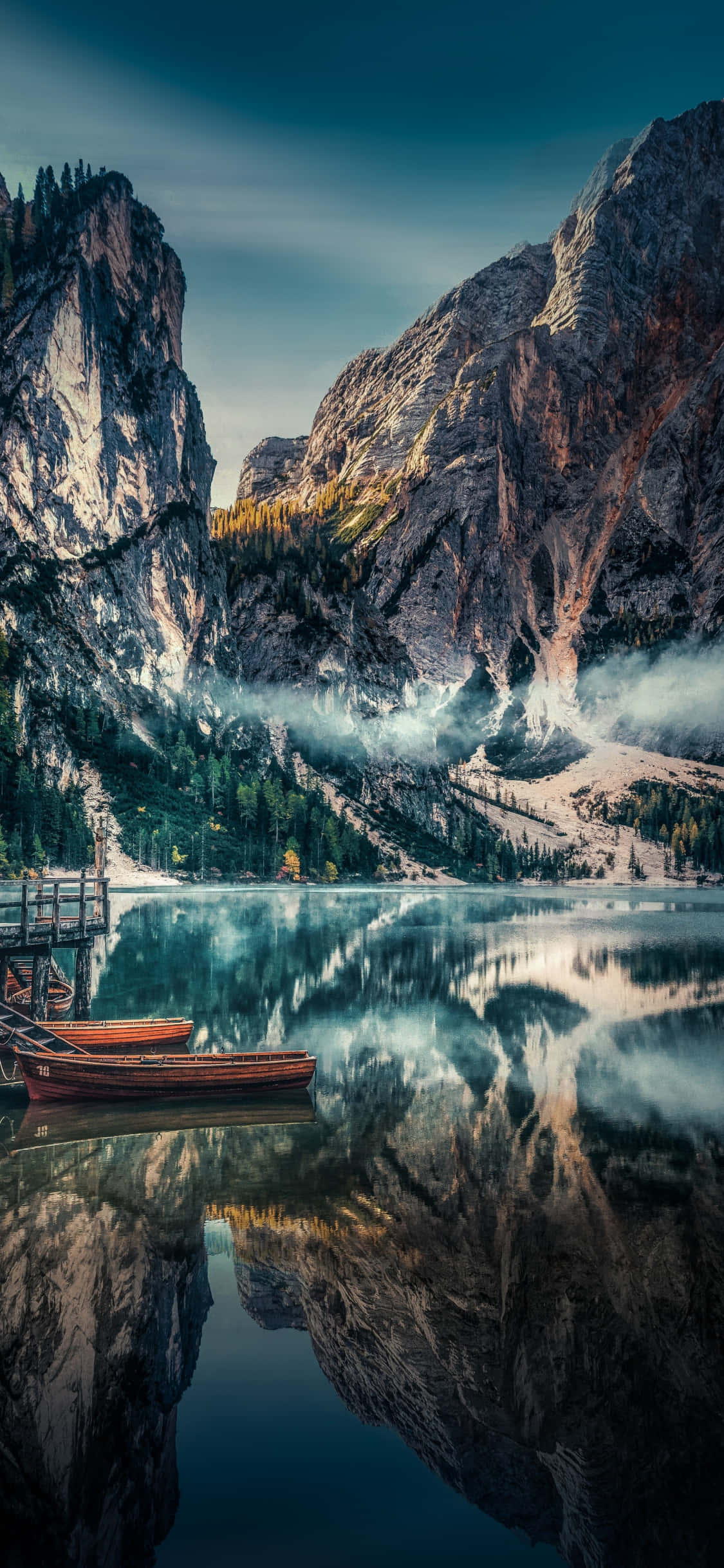 iPhone X Nature 桌布 壁紙 — 一個寧靜的山中湖，在暮色天空下映照著高聳的懸崖和霧氣繚繞的山峰，兩艘木船停泊在碼頭。