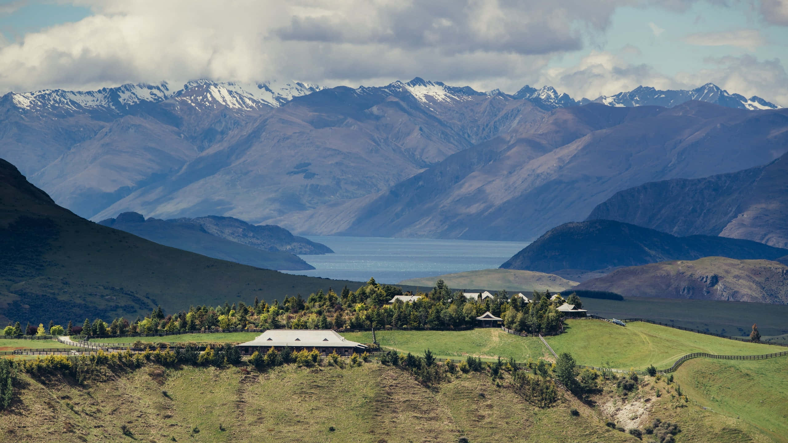 New Zealand 桌布 壁紙 — 一個寧靜的山谷，農舍坐落在起伏的綠色山丘之間，俯瞰著平靜的藍色湖泊和遠處白雪覆蓋的山脈，天空部分多雲。