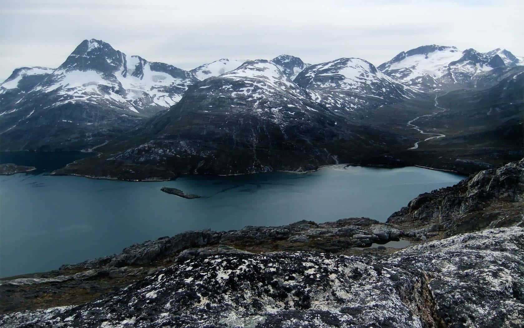 Greenland 桌布 壁紙 — 一個寧靜的山間湖泊，坐落在積雪的山峰之間，天空多雲，一條蜿蜒的道路穿過山谷。