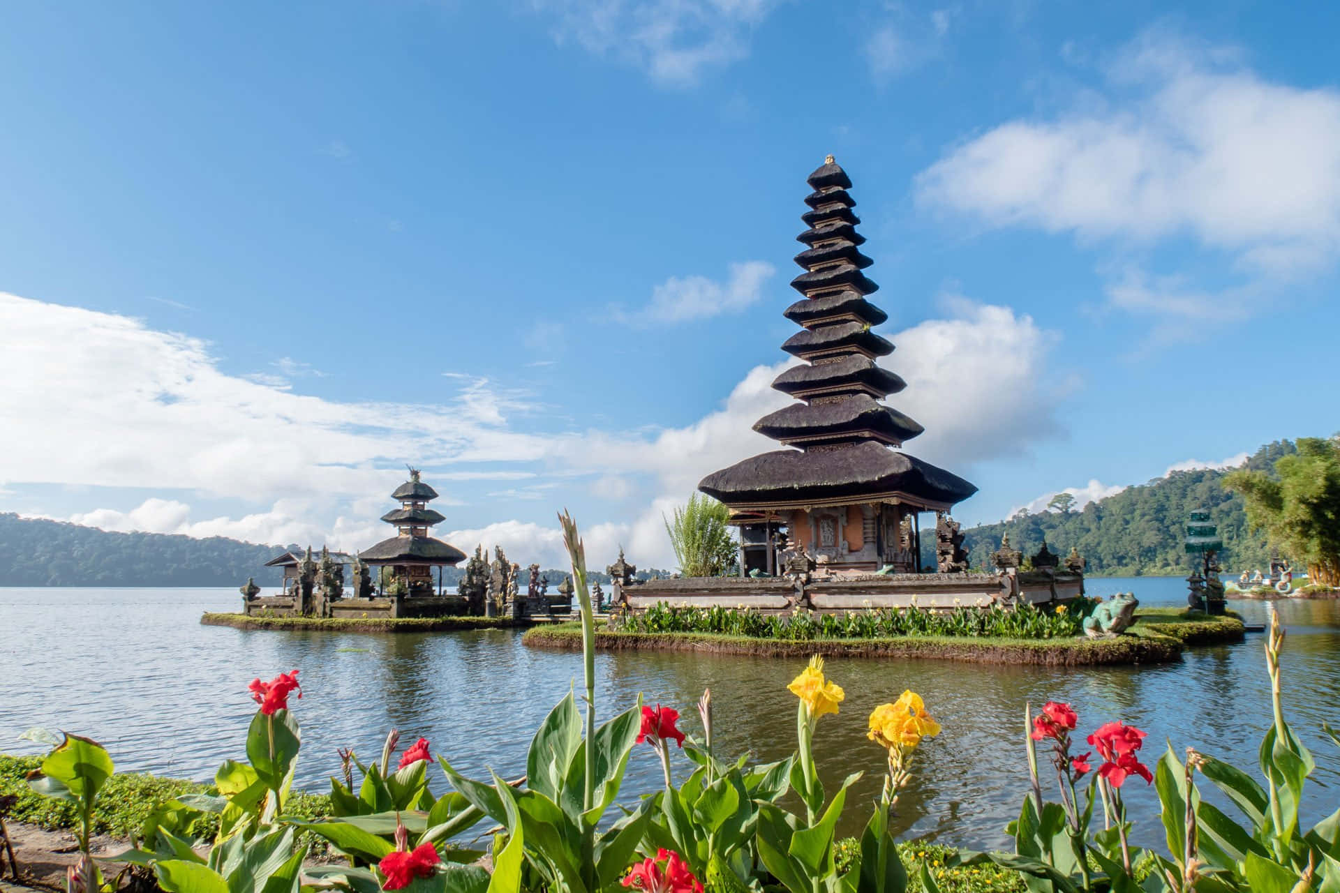 Bali 桌布 壁紙 — 一個寧靜的峇里島寺廟建築群，擁有層疊的屋頂，優雅地坐落在平靜的湖面上，周圍是鬱鬱蔥蔥的綠色山丘，前景是鮮豔的紅色和黃色花朵，在明亮的藍天下。