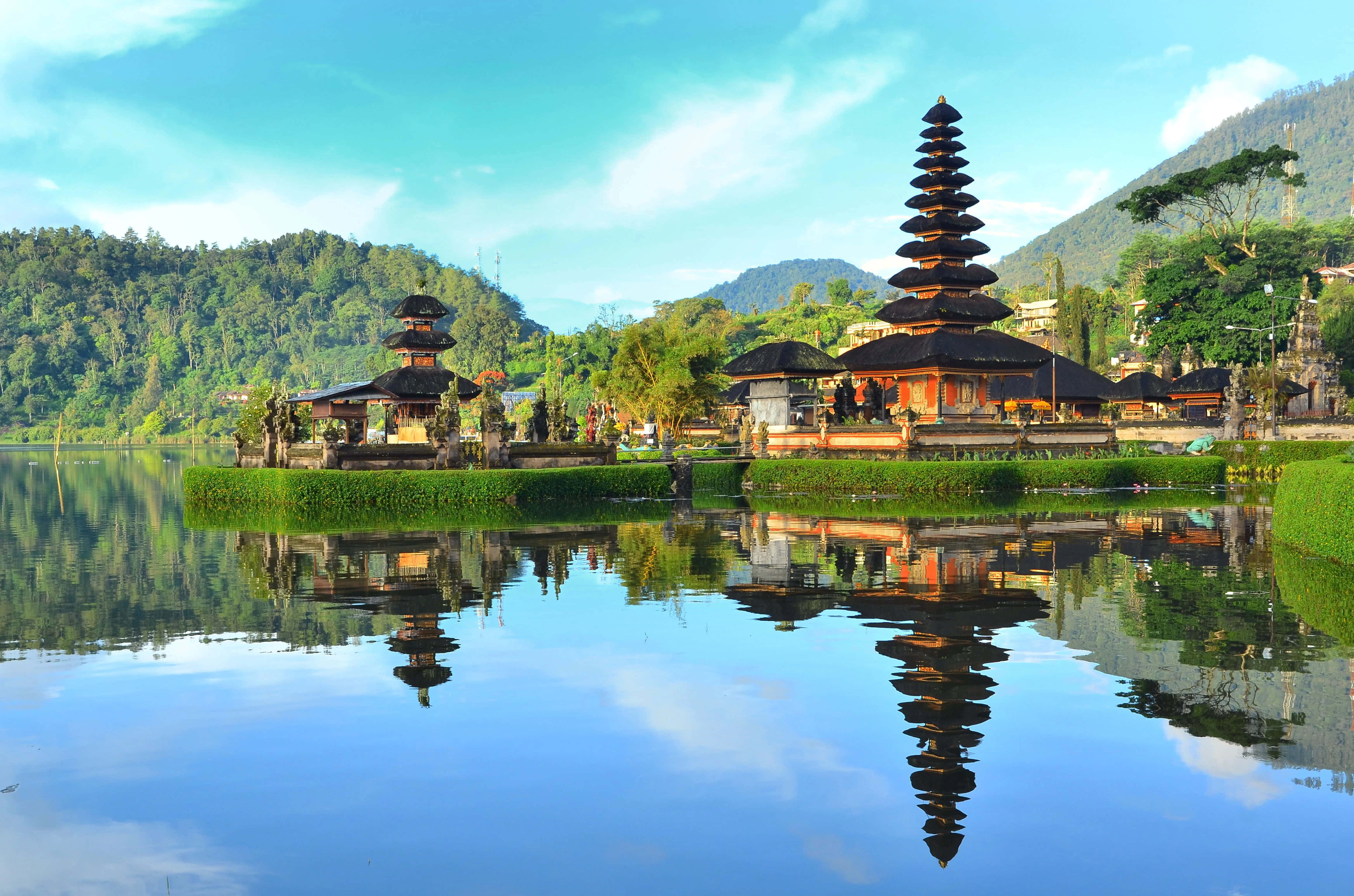 Bali 桌布 壁紙 — 一個寧靜的峇里島寺廟群，擁有層疊屋頂，完美倒映在平靜的藍綠色水域中，四周環繞著蒼翠的綠色山丘，上方是明亮的藍天。