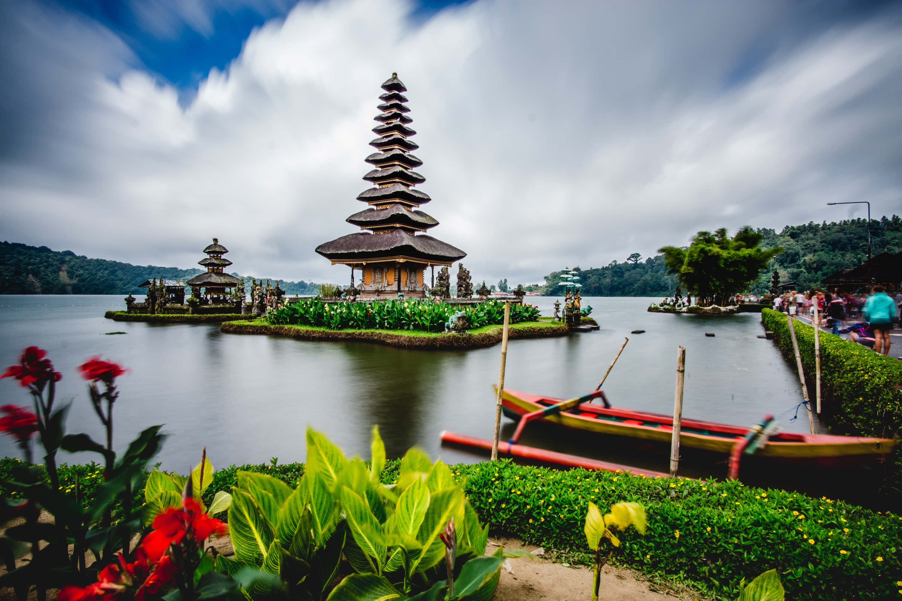 Indonesia 桌布 壁紙 — 一個寧靜的峇里島寺廟群漂浮在平靜的湖面上，周圍環繞著茂密的綠色植物和鮮豔的紅花，天空多雲。