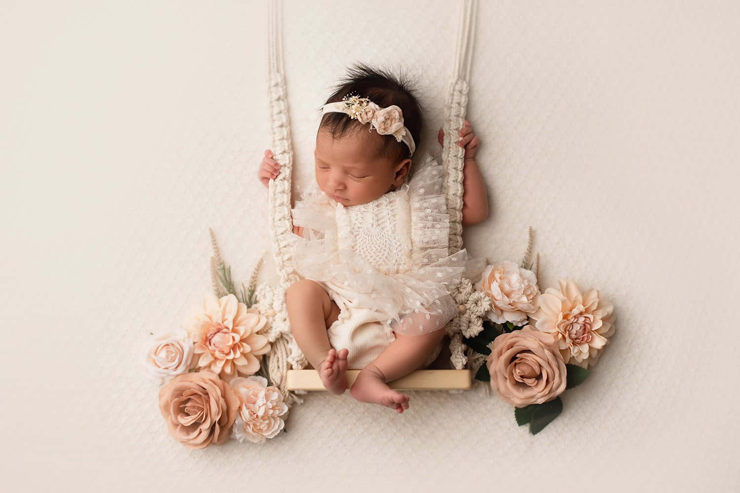 Newborn Baby 桌布 壁紙 — 一個寧靜的新生嬰兒在流蘇編織吊床上安睡，周圍環繞著柔和的蜜桃色和奶油色花朵，背景是帶有紋理的白色。