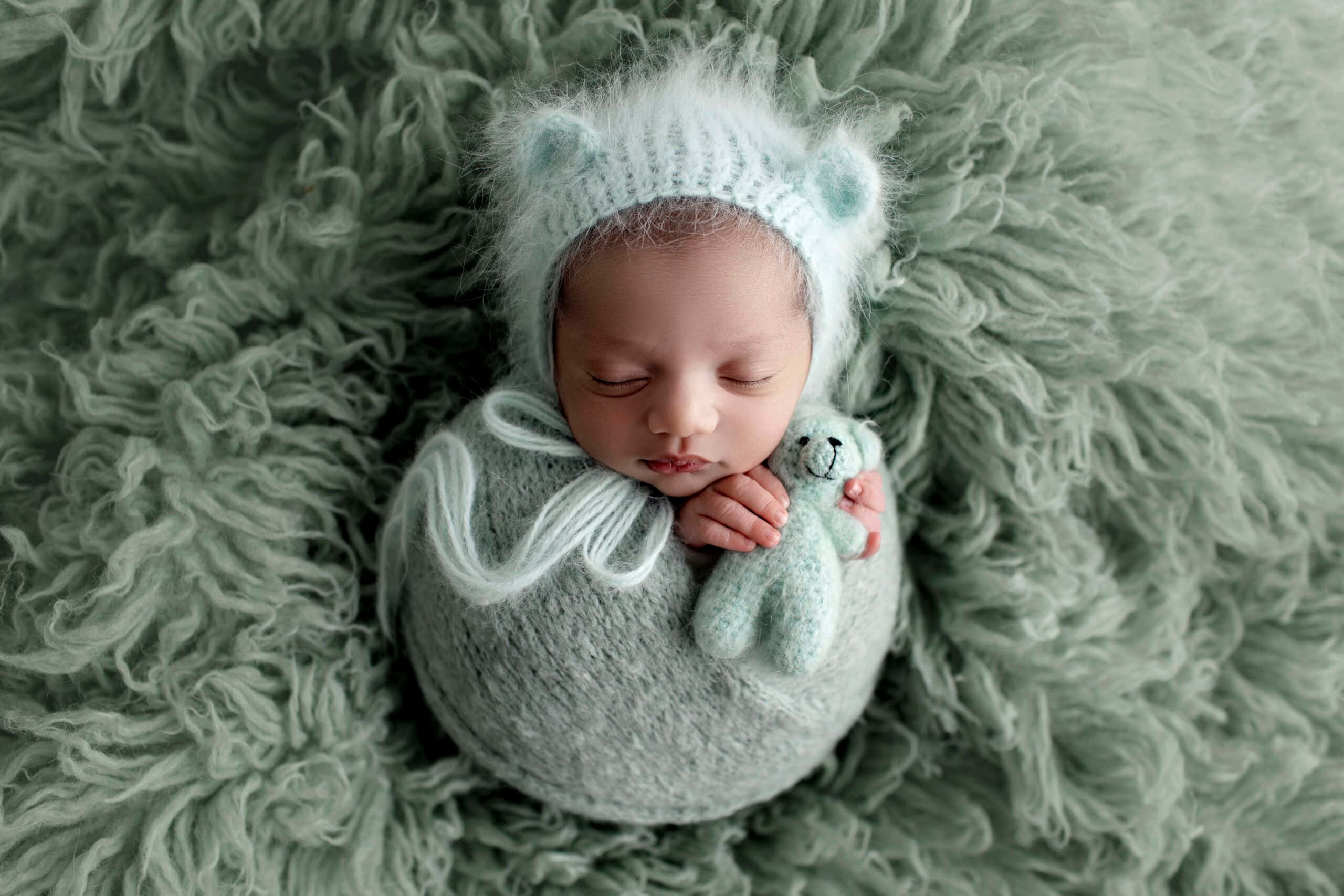 Newborn Baby 桌布 壁紙 — 一個寧靜的新生嬰兒被柔軟的薄荷綠色布料包裹著，在毛絨地毯上抱著一隻小編織熊，安詳地睡著。